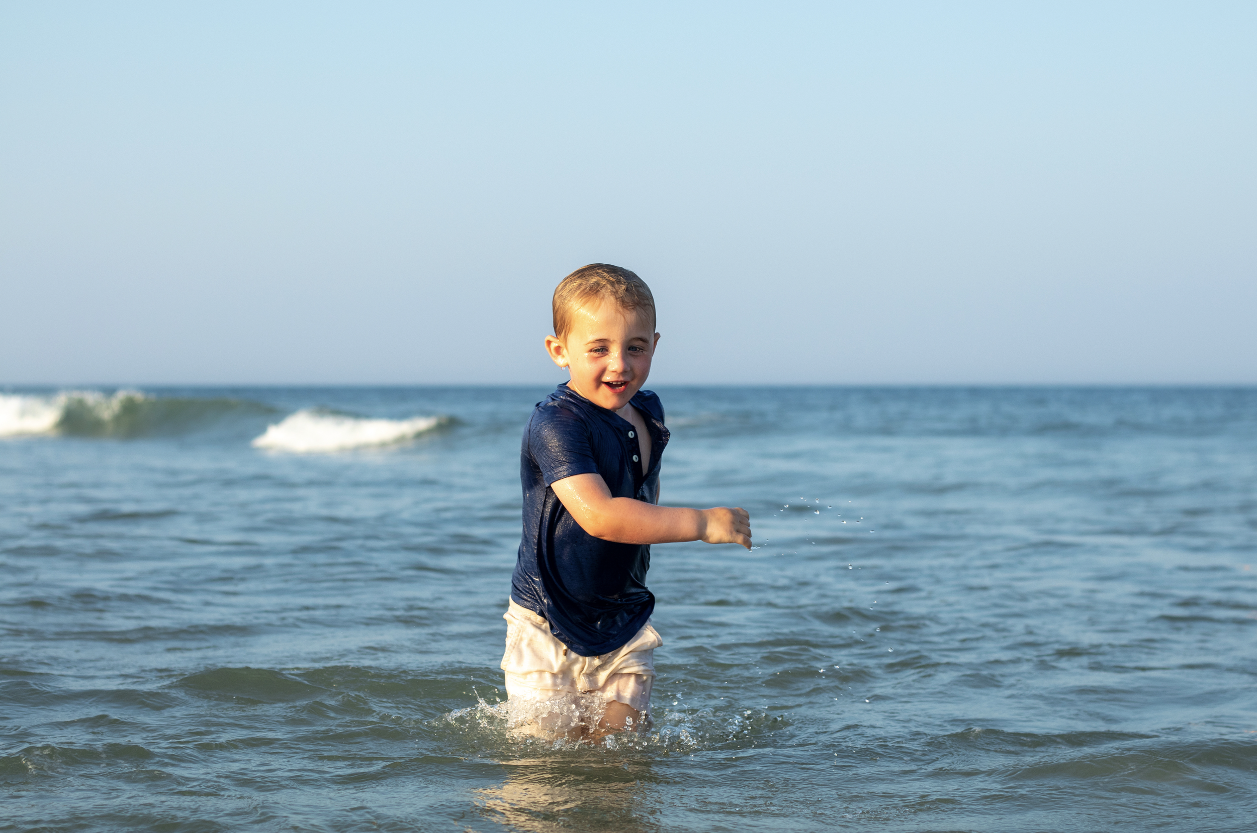 Young boy playing in the ocean water at the beach, smiling and splashing with waves in the background. Lavallette, Ocean County, New Jersey Photographer. Family session with In Water add on