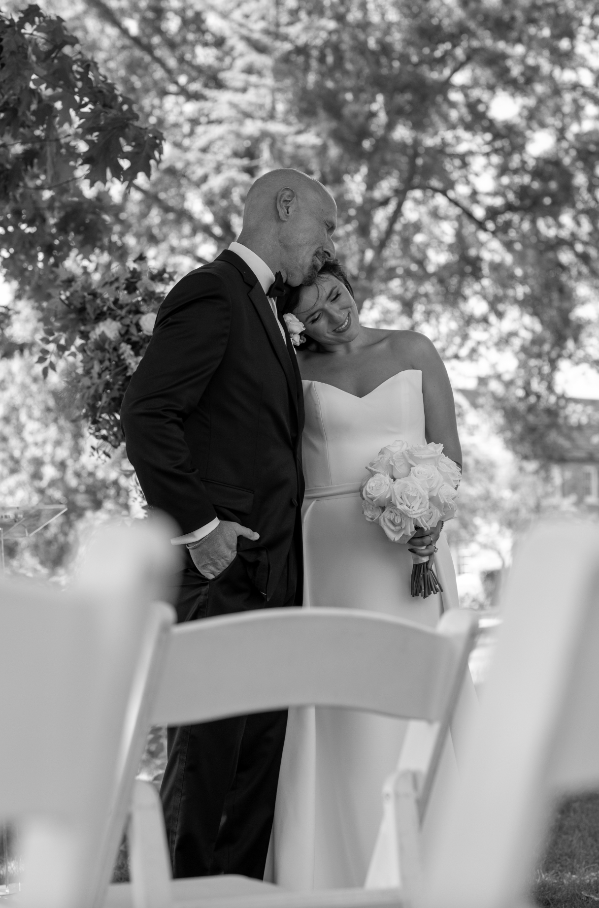 Black and white photo of a bride and an older man, possibly her father, standing outdoors under trees. The bride holds a bouquet of roses and smiling, with her head resting on the man's shoulder, both dressed in formal attire for a wedding.
