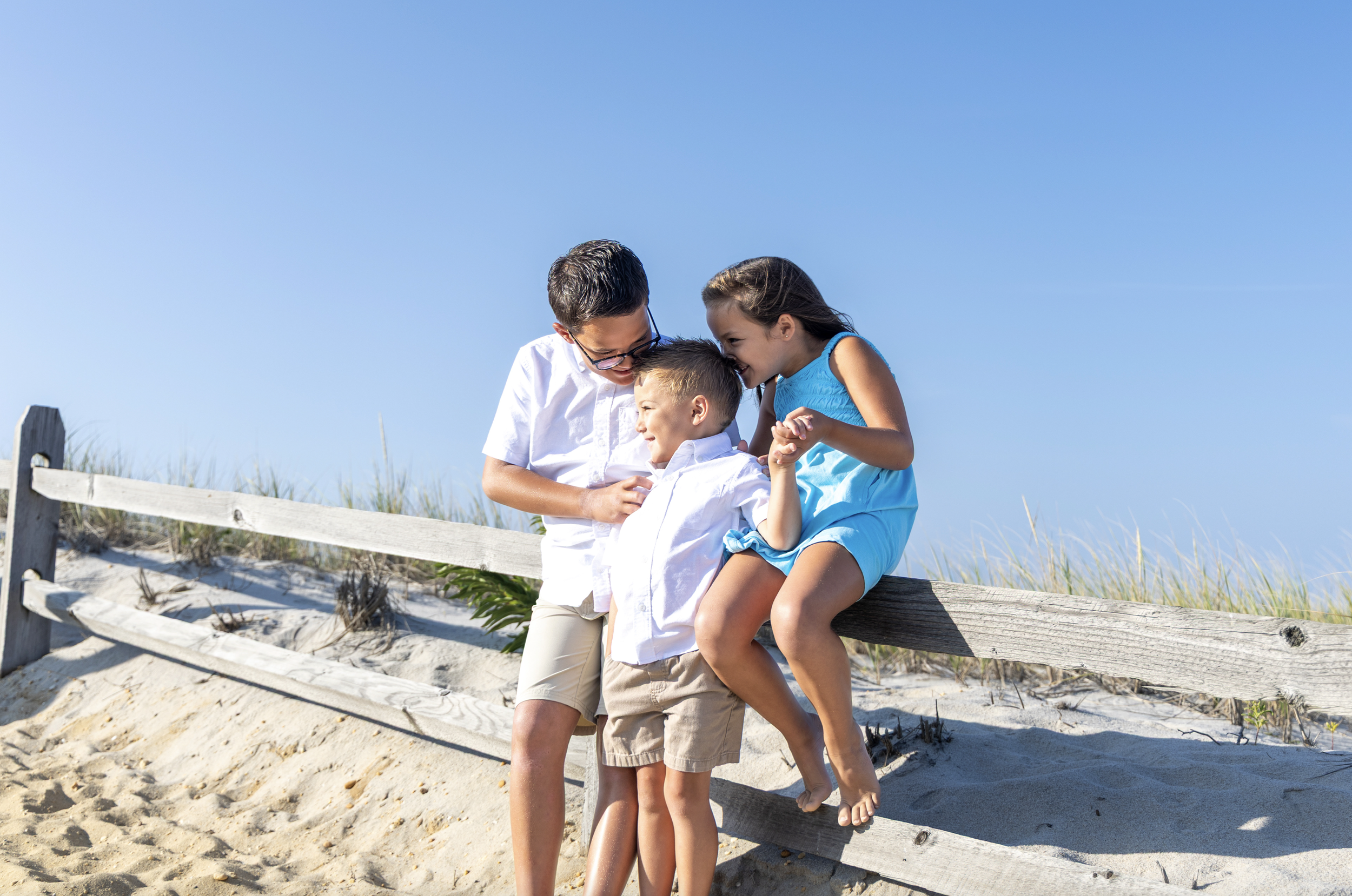 Three children playing and laughing together on a wooden fence at the beach on a sunny day. New Jersey beach photography