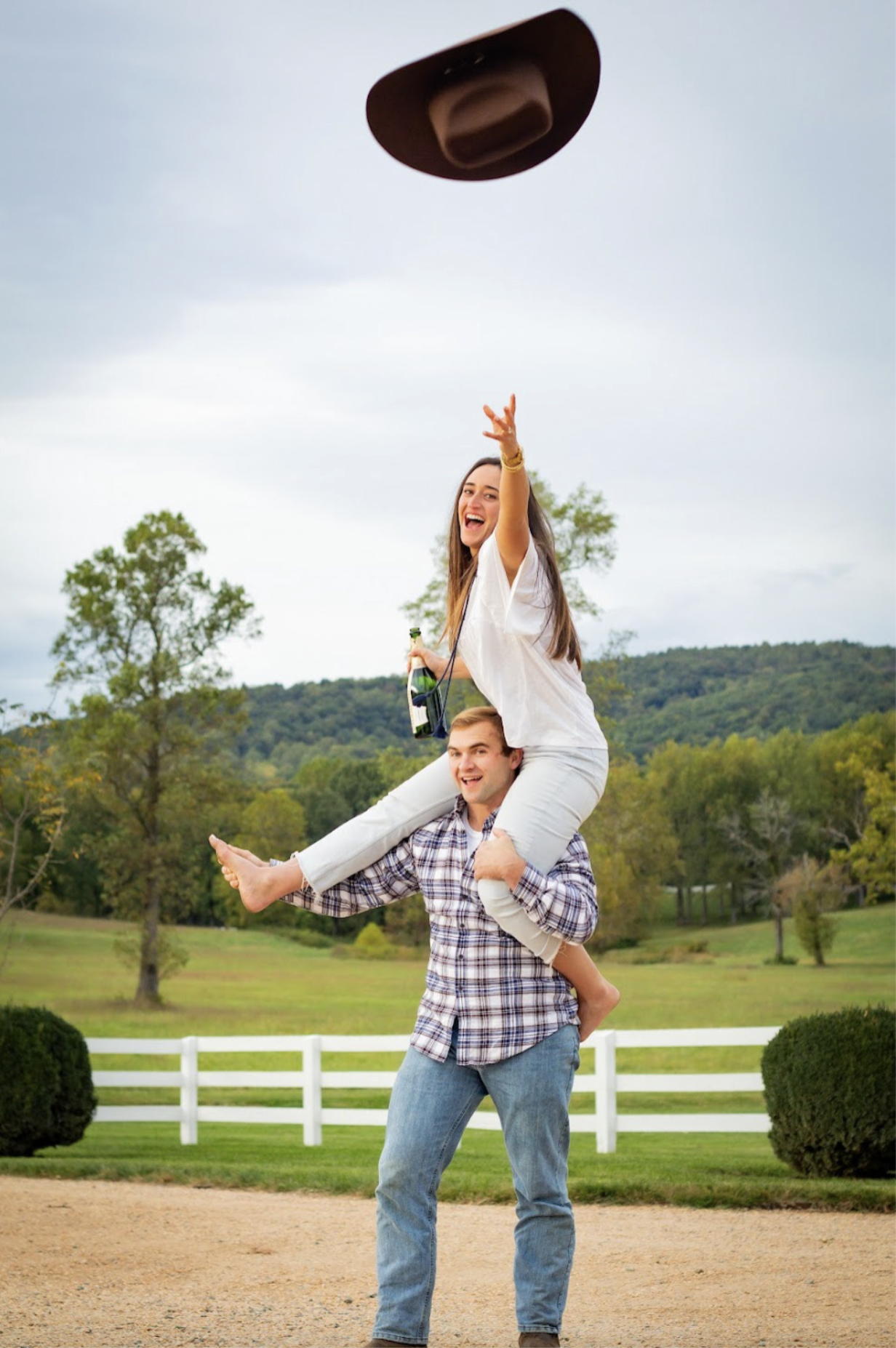 A young woman sitting on a young man's shoulders, throwing a hat into the air and smiling, outdoors in a rural area with trees and green hills in the background. rustic engagement photographer