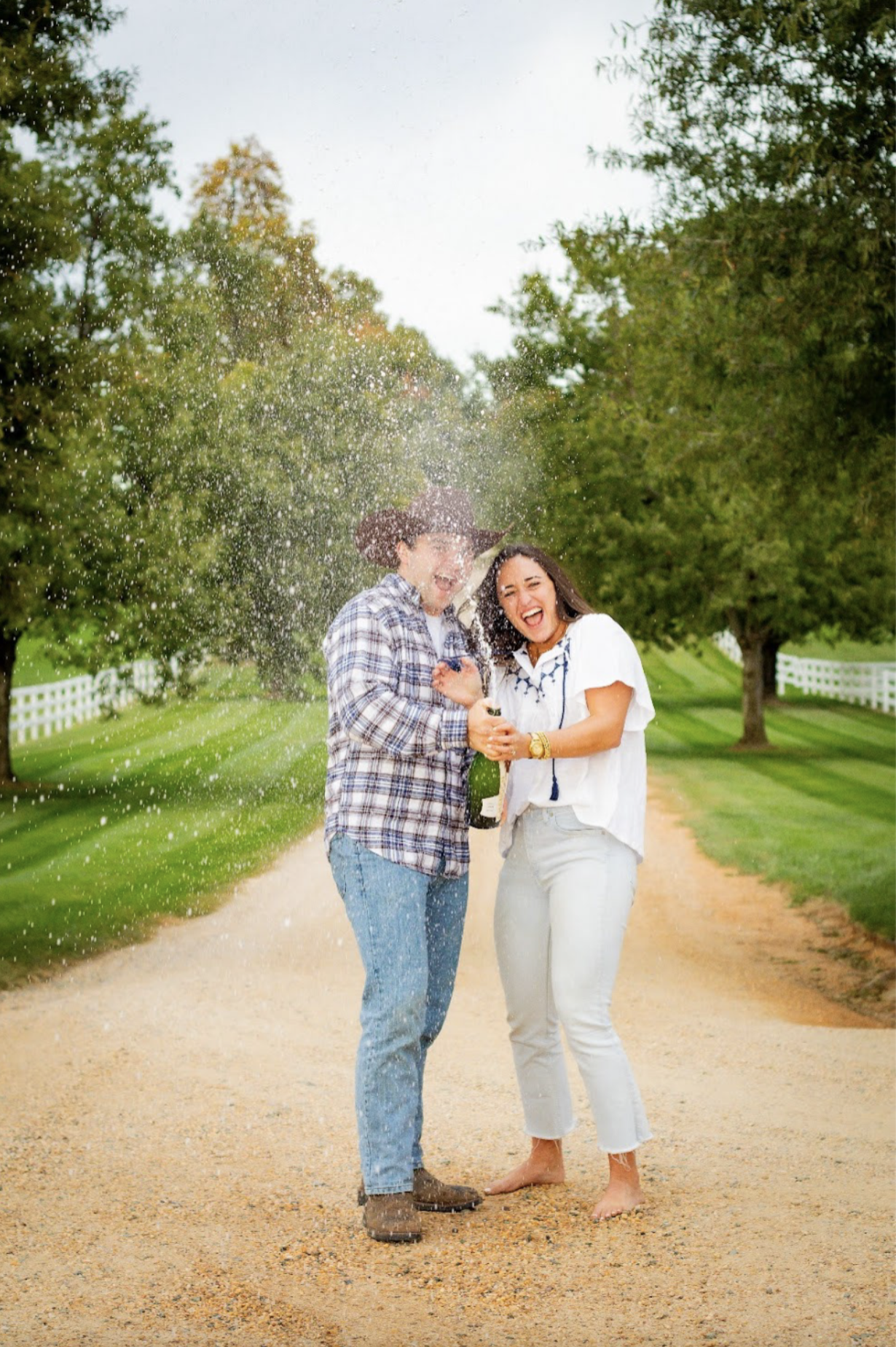 A man and woman celebrating on a country road, with the woman opening a bottle of champagne and both laughing, as champagne sprays in the air. Rustic engagement session. Engagement Photography