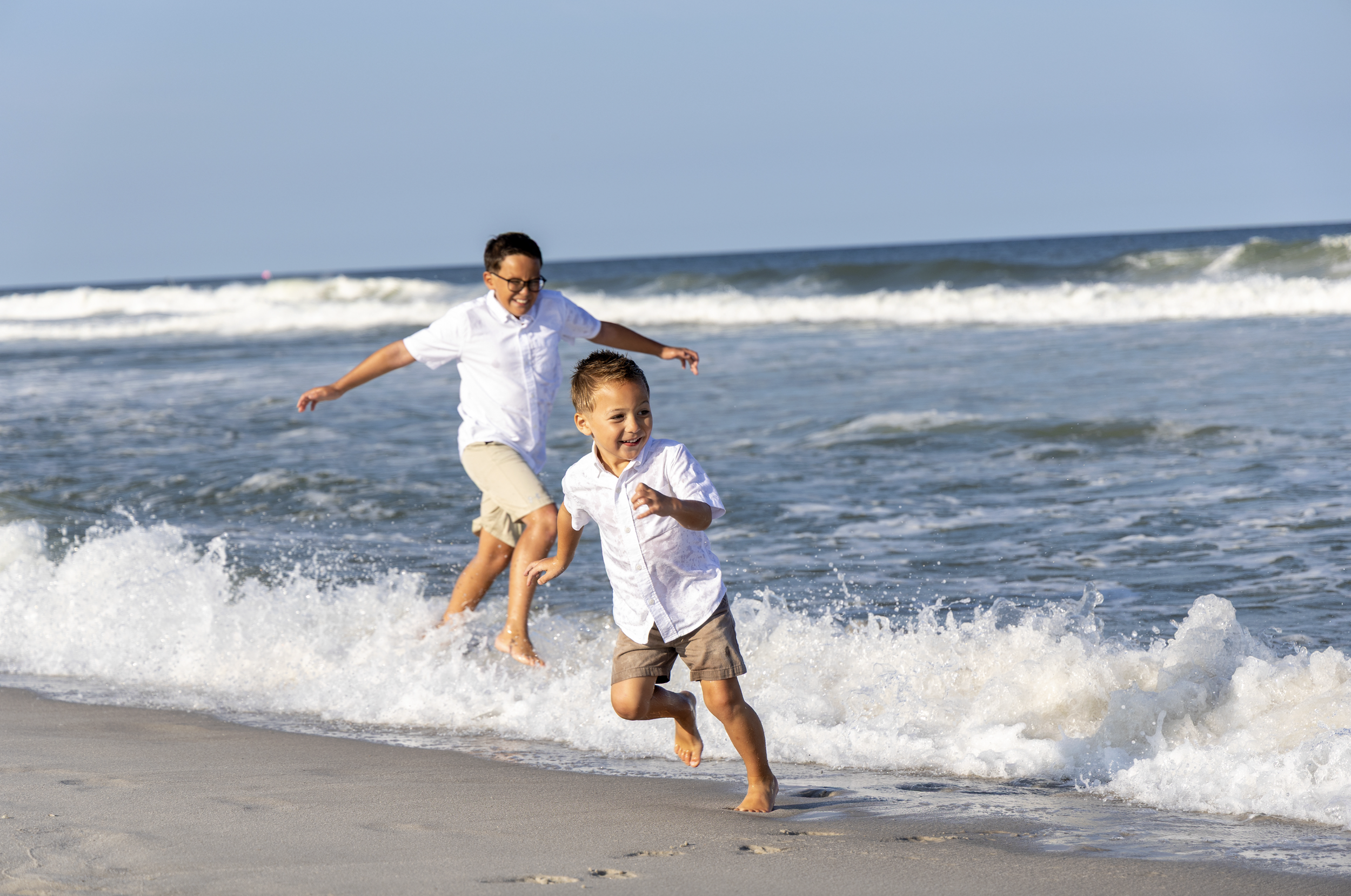 Two young boys in white shirts and shorts playing in the ocean waves at the beach, smiling and running. In Water add on Beach photography, new jersey