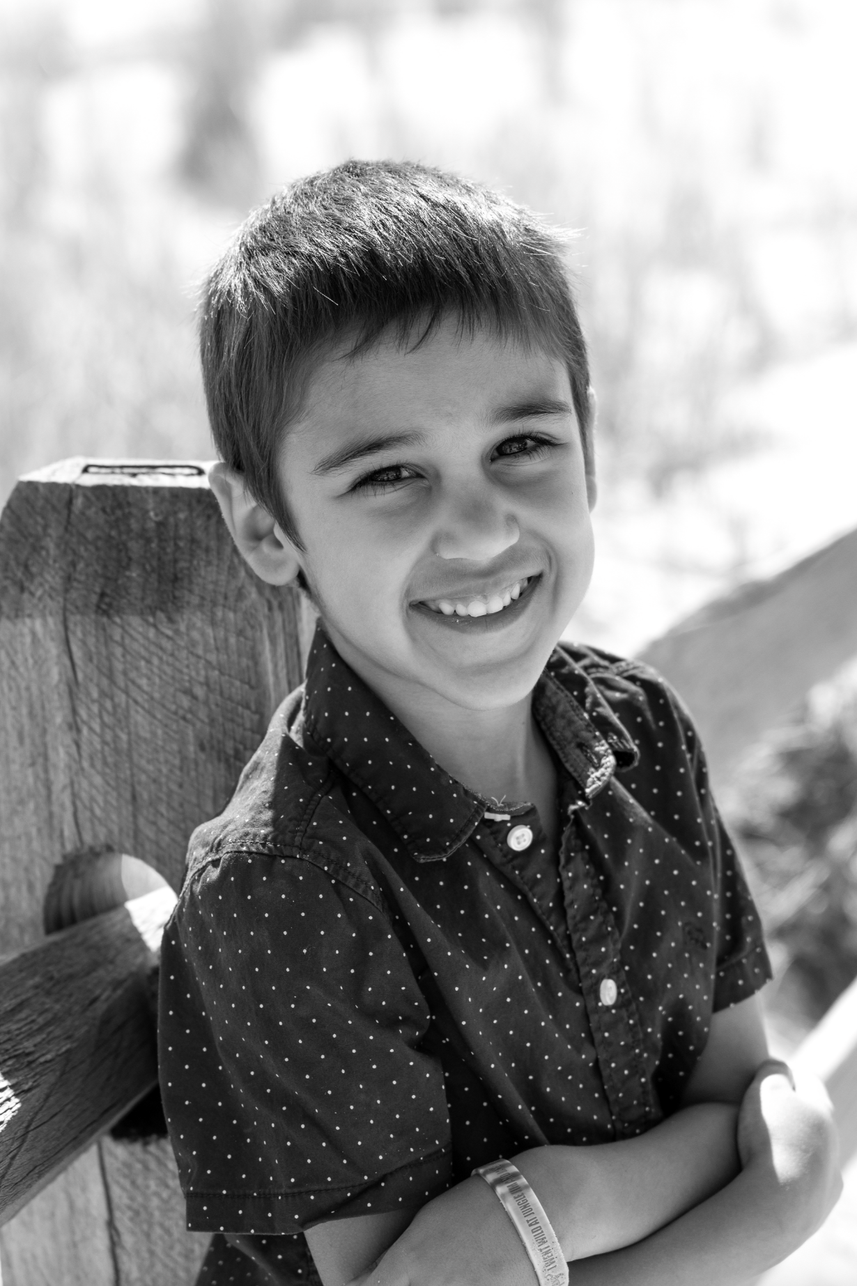 A smiling young boy with short dark hair, wearing a dark polka dot shirt, sitting on a wooden bench outdoors.
