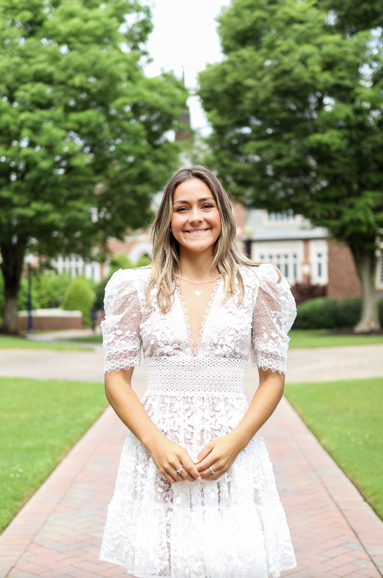 Young woman smiling in white lace dress standing on a brick sidewalk in a park with green trees and buildings in the background. New Jersey graduation