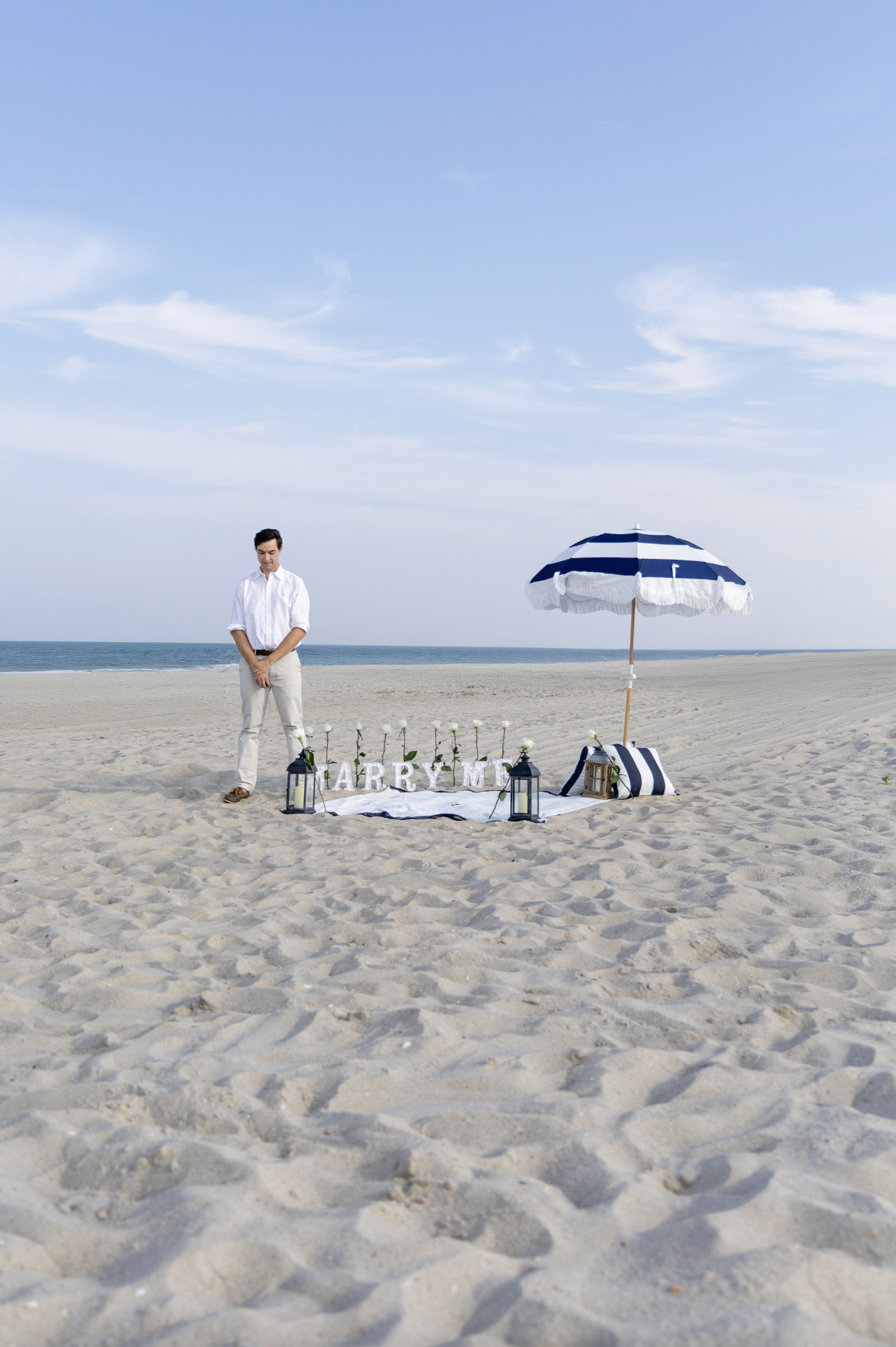 A man standing on a sandy beach next to wedding decor including a white cloth with the word "Marry Me" and flowers, a striped umbrella, lanterns, and a bag, with the ocean and blue sky in the background. Ocean County, New Jersey Photographer
