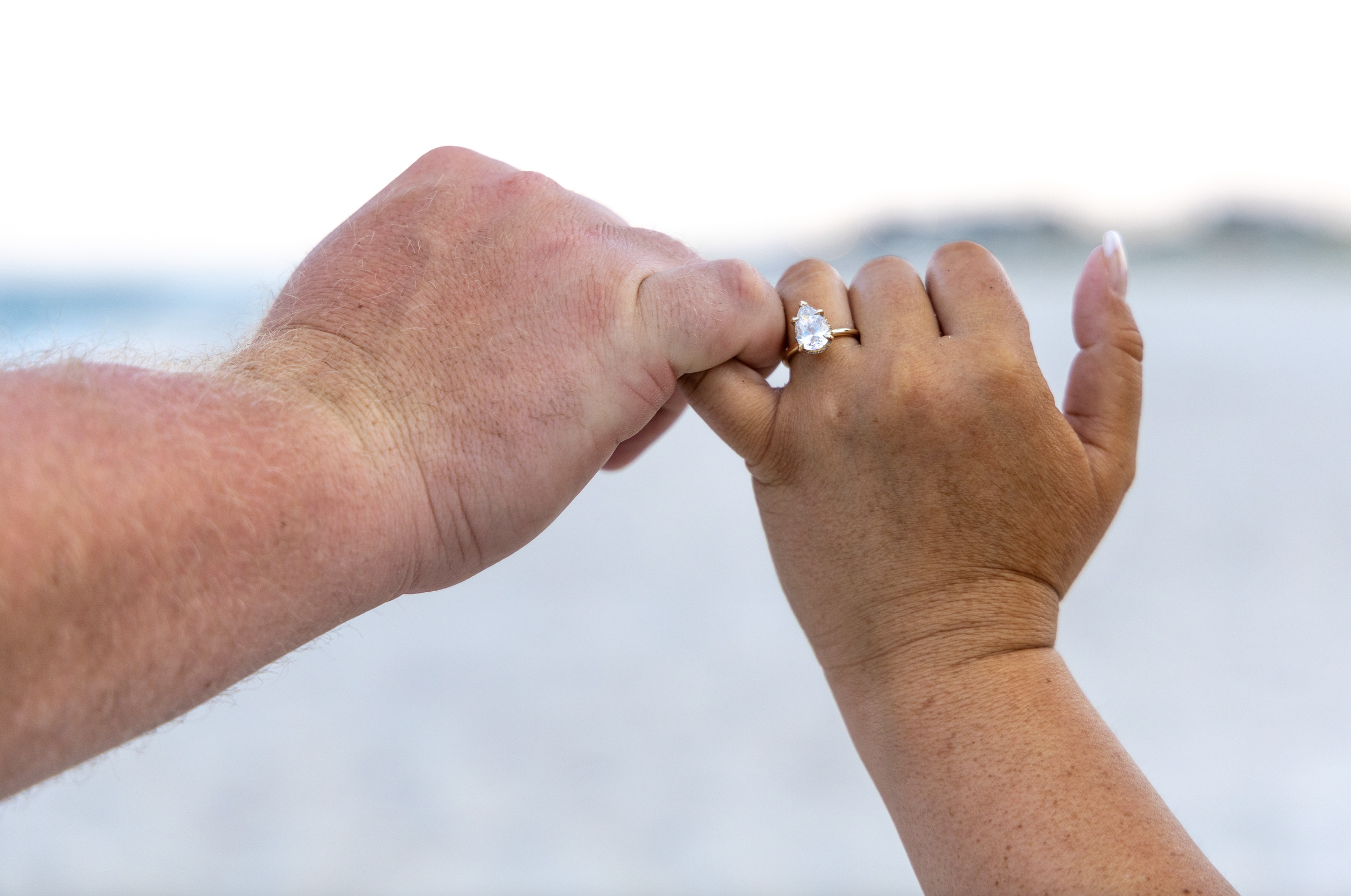 Two hands holding each other, one hand with a diamond ring, against a blurred ocean background. Ocean County, New Jersey Photography