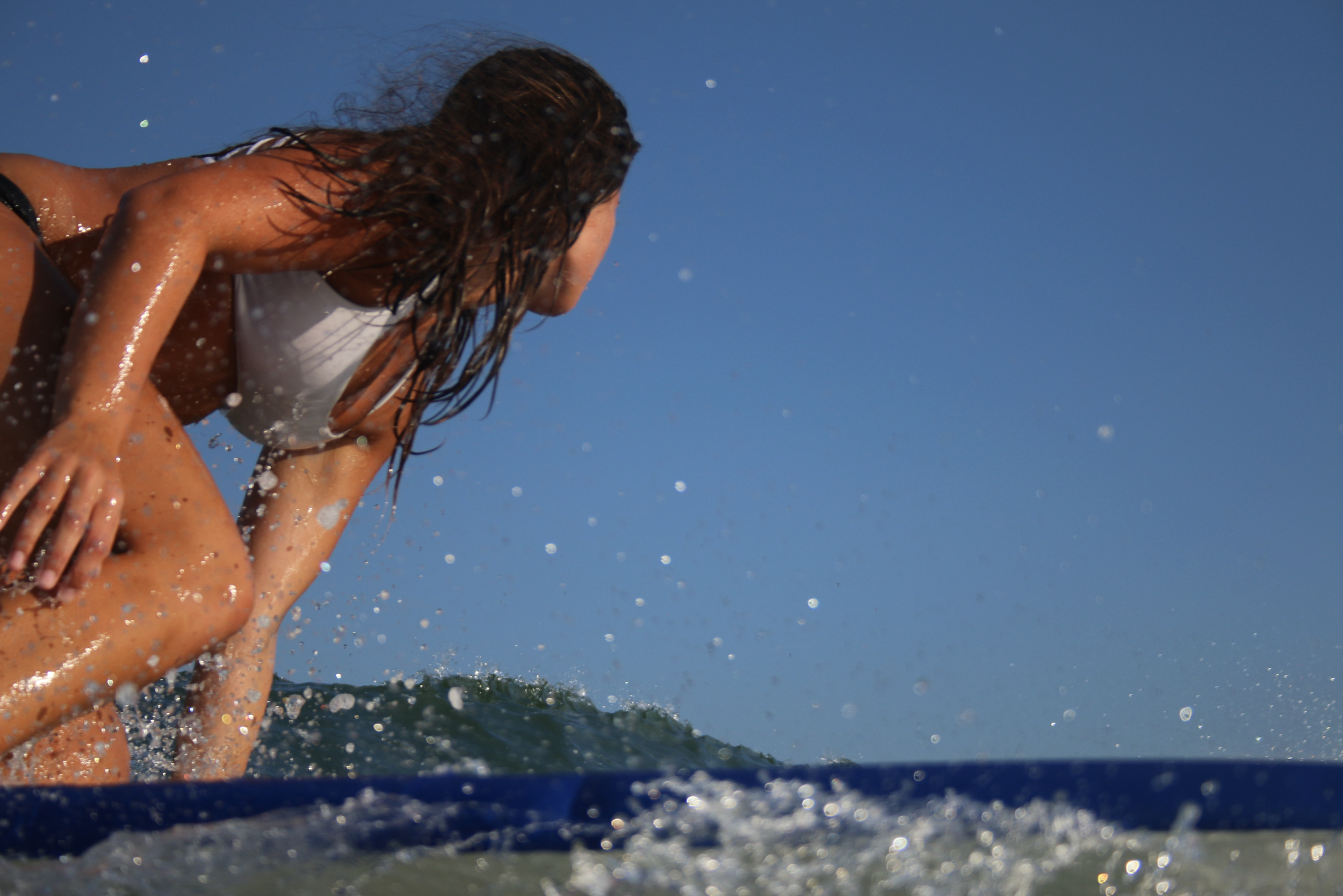 A woman with wet hair in a white swimsuit leaning over a person on a surfboard in the ocean, blue sky in the background.