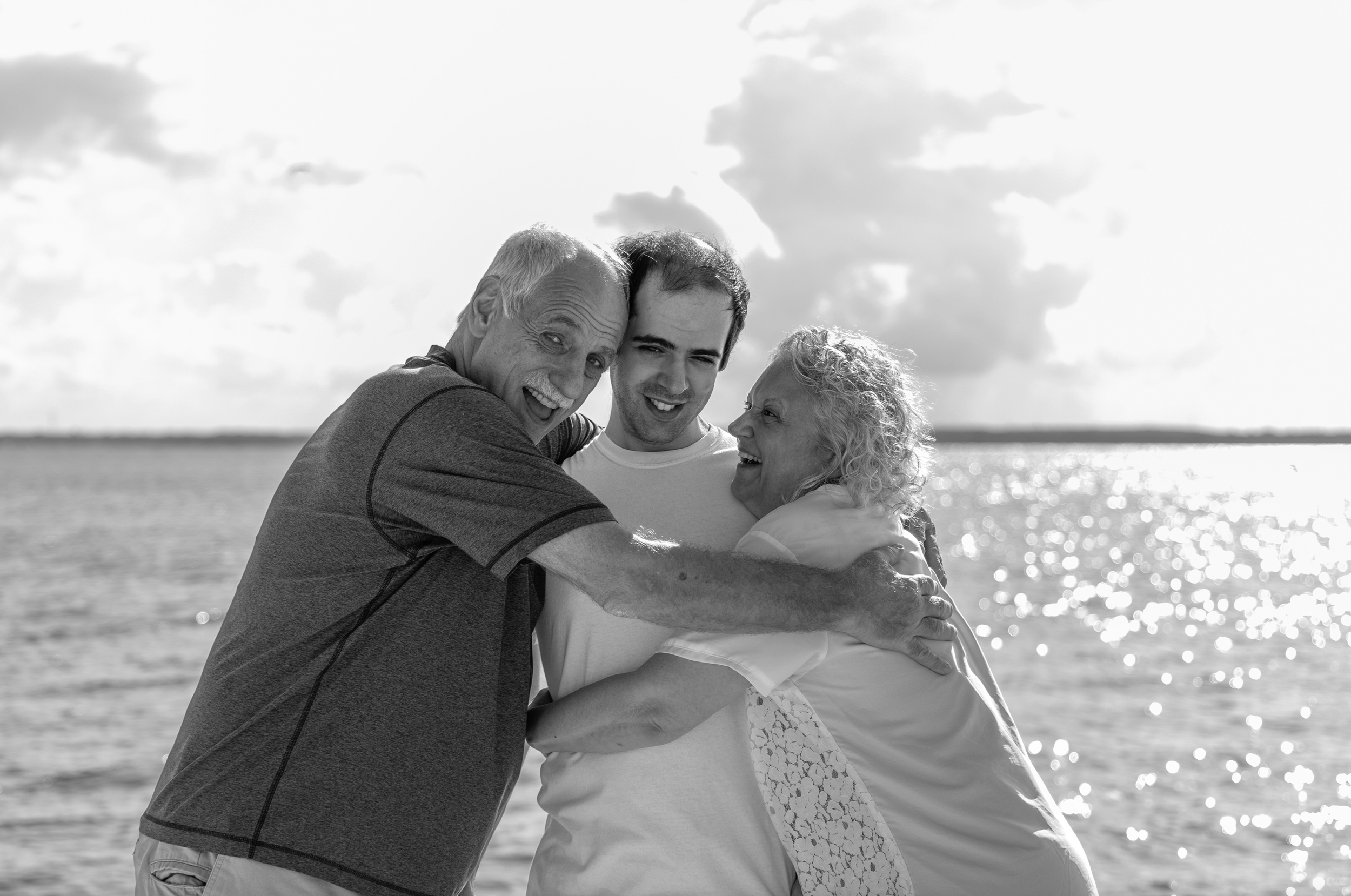 Four people, two elderly and two young adults, hugging and smiling on a beach with water and cloudy sky in the background. Adult Family Photos, Ocean County New Jersey