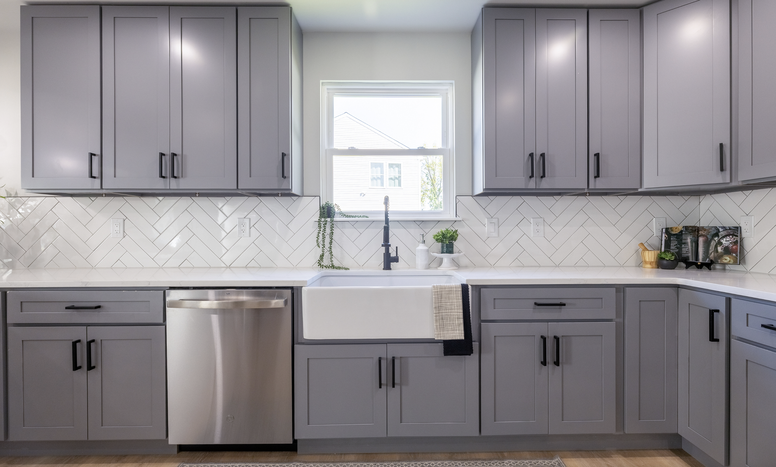 Kitchen with gray cabinets, white countertop, and white herringbone tiled backsplash. A single window above a farmhouse sink with a black faucet. Small plants and a soap dispenser on the counter near the window. To the right, a small display with a cookbook, plant, and decorative items.