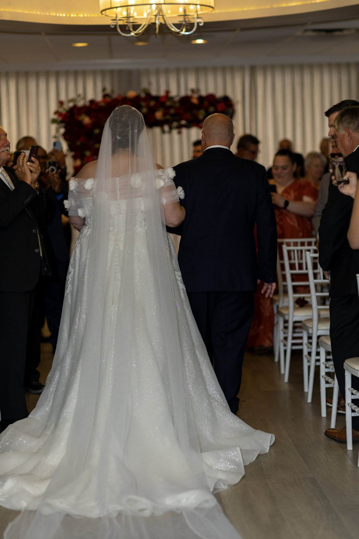 Bride and groom walking down the aisle at their wedding, surrounded by seated guests and photographers, with floral decorations in the background.