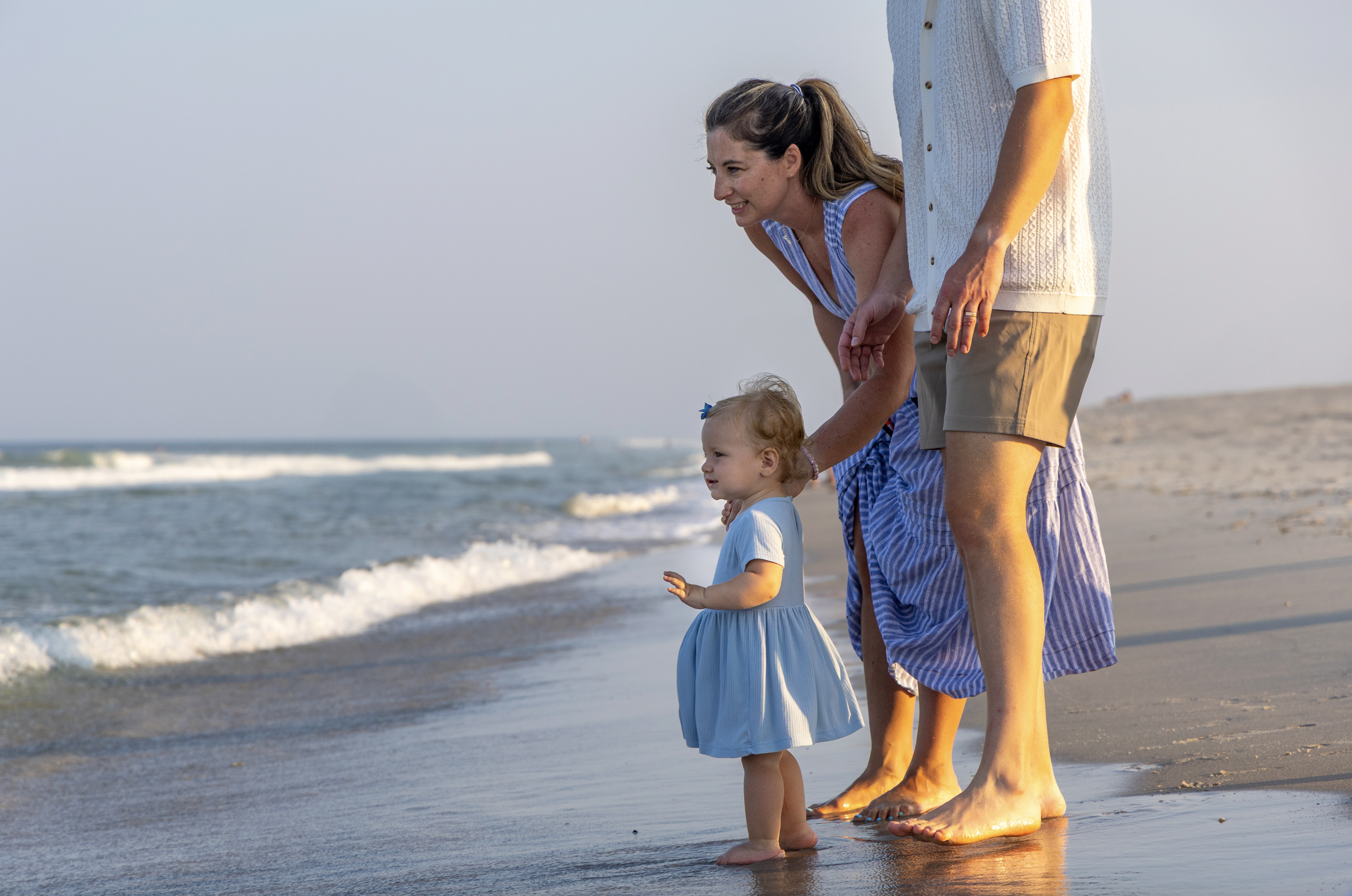 A family of three, including a young girl in a blue dress, stands barefoot on the beach with waves at sunset, smiling and looking at the ocean. Lavallette, New Jersey Family photographer
