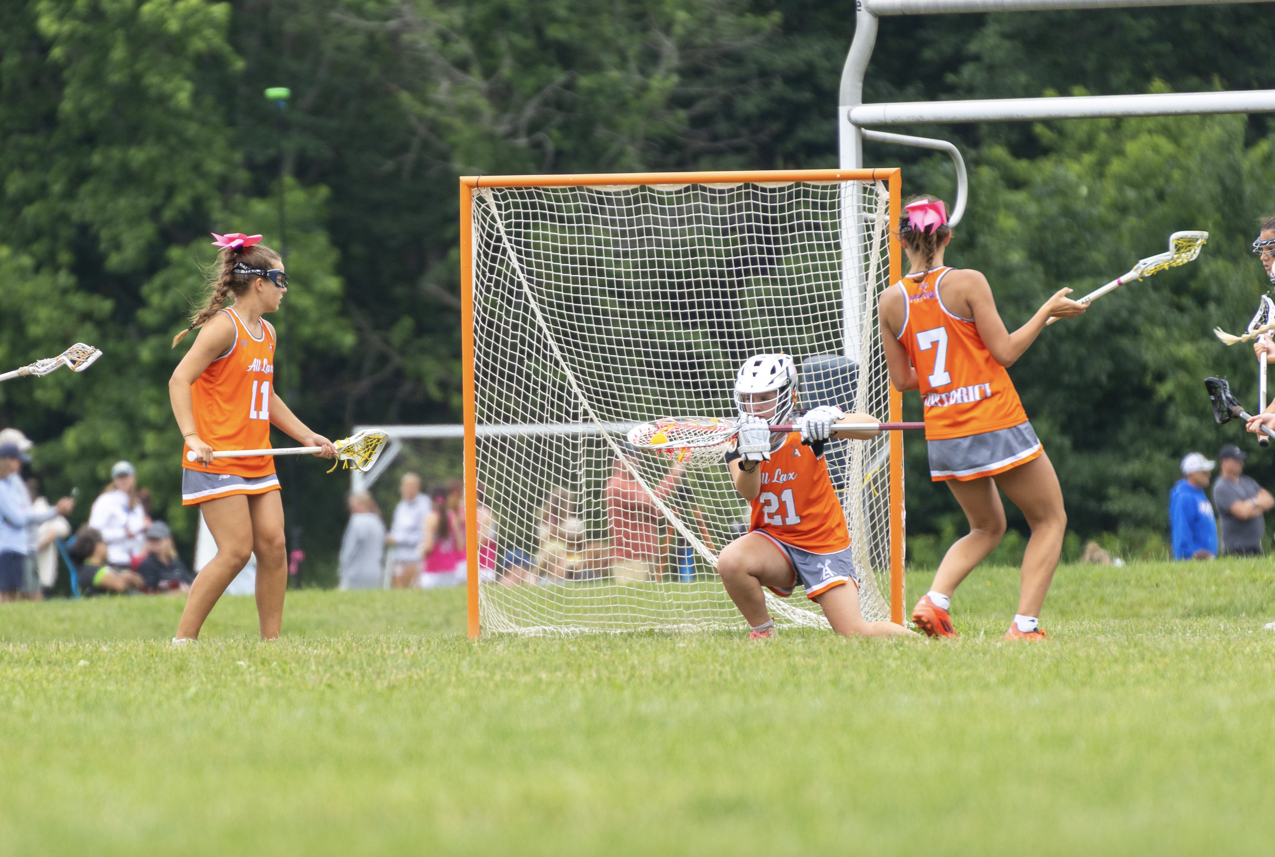 Women playing lacrosse on a grassy field, with one goalie in a helmet kneeling in front of the goal, two players wearing orange jerseys with pink bows and lacrosse sticks, and spectators in the background.