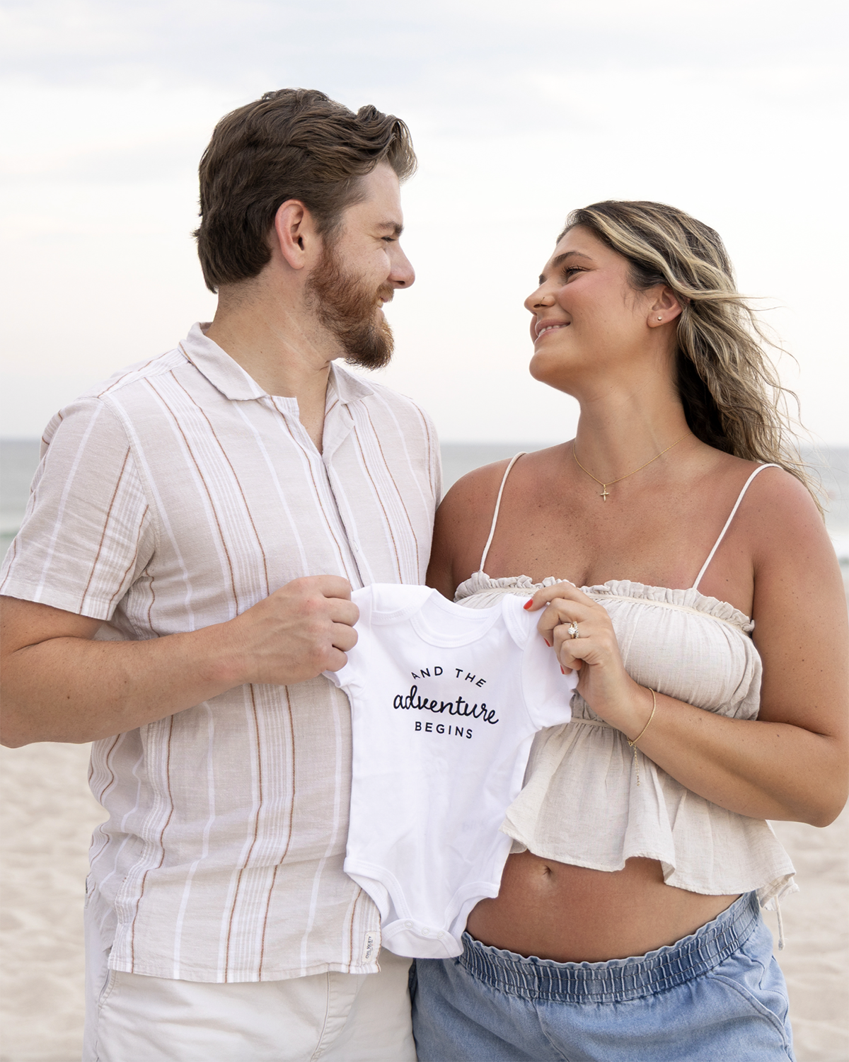 A young couple standing on a beach, holding a baby onesie that says 'and the adventure begins,' and smiling at each other. Beach Maternity Sessions New Jersey