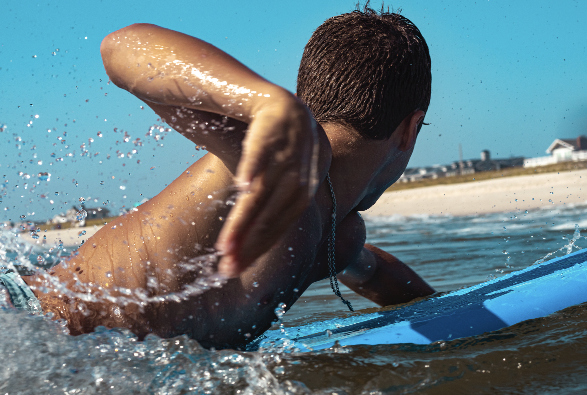 A man with short dark hair surfing on a blue surfboard in the ocean, viewed from behind, with a sandy beach and buildings in the background.