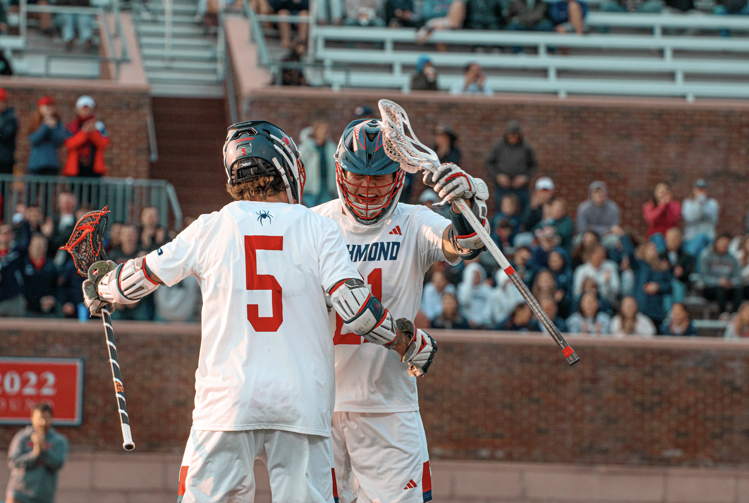 Two lacrosse players wearing white jerseys with red numbers 5 and 21 celebrate on the field, with a crowd of spectators in the stands behind them.