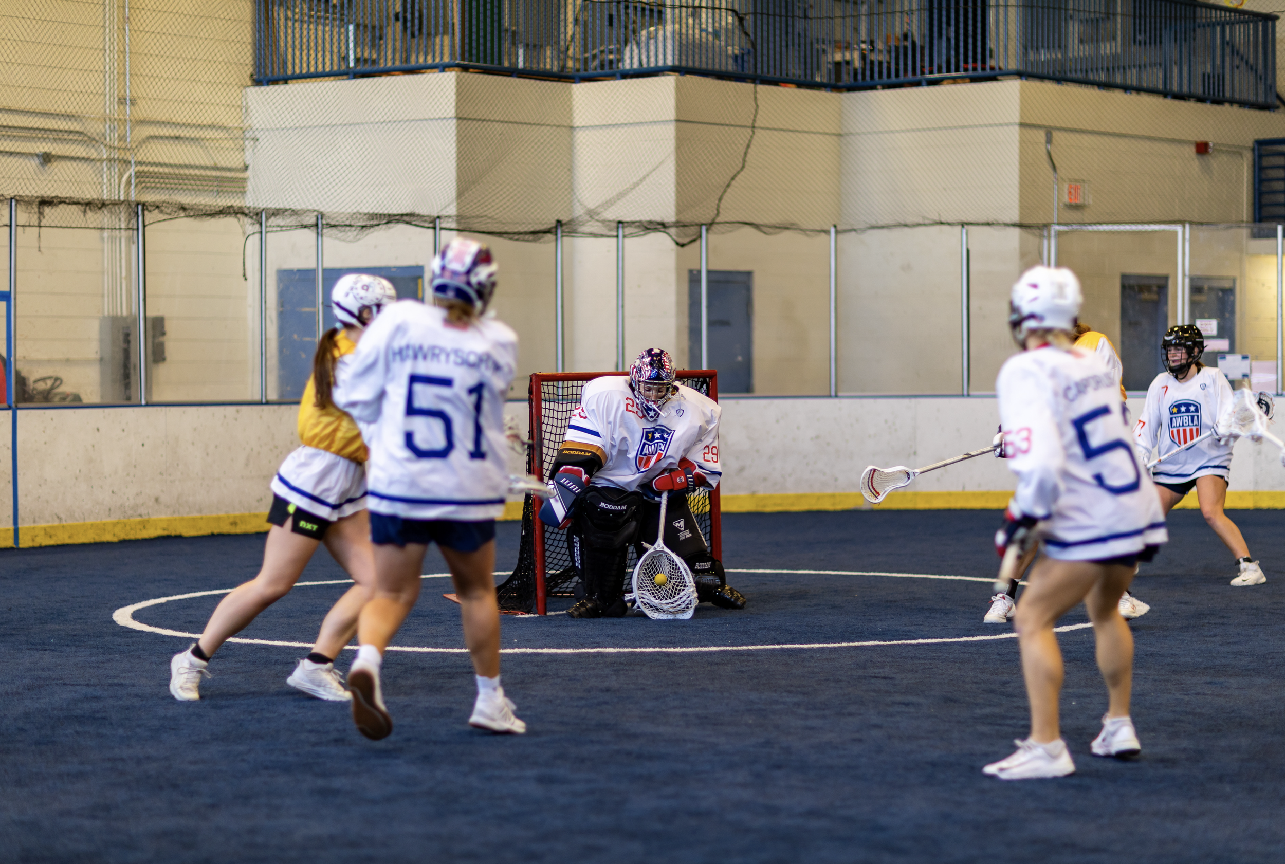 Women playing indoor lacrosse game, with goalie in front of net and several players in white jerseys and helmets around her.