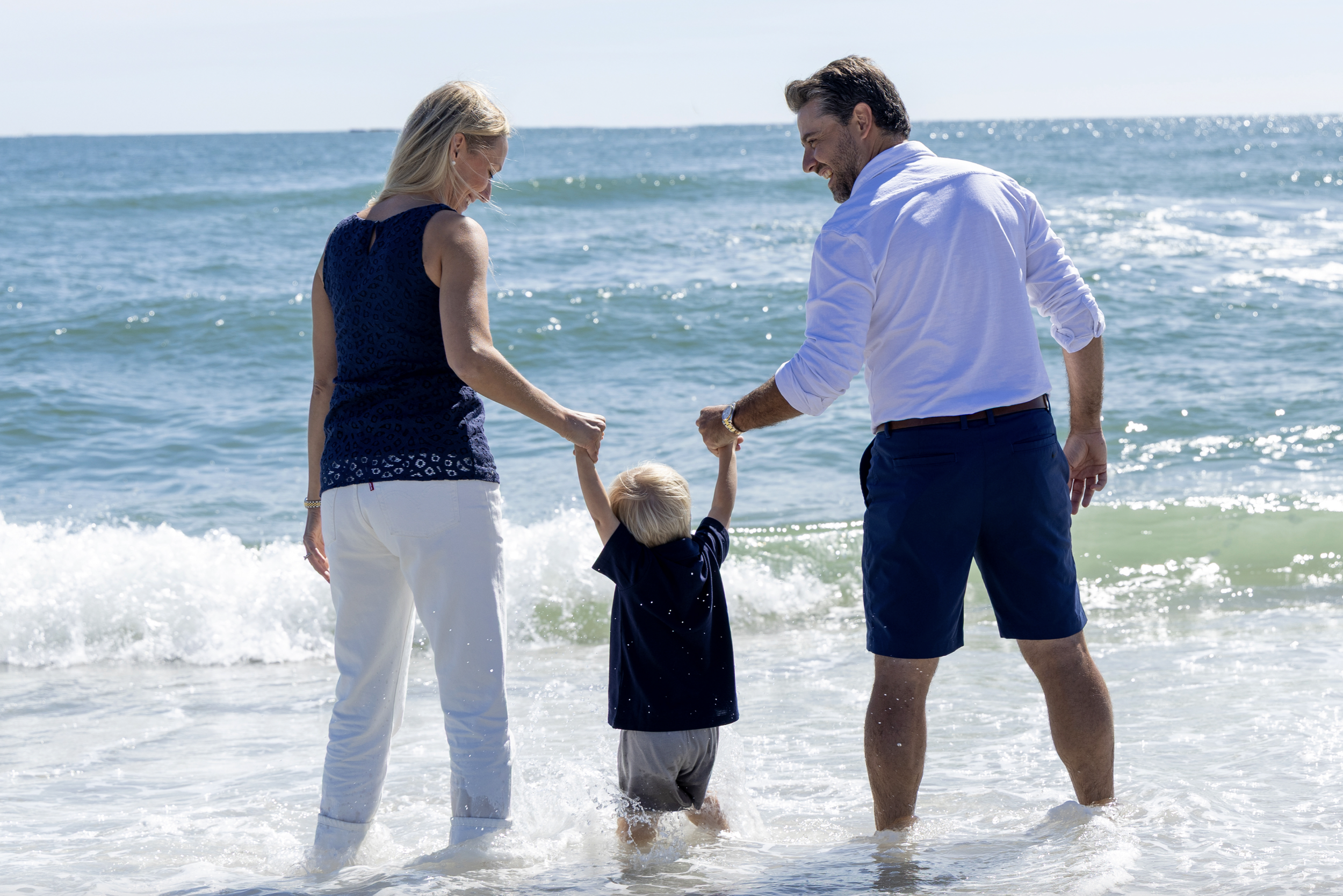 A family of three on the beach, a woman, a man, and a young child, holding hands and walking in the shallow water near the shoreline with waves and ocean in the background. Monmouth County, New Jersey Photography