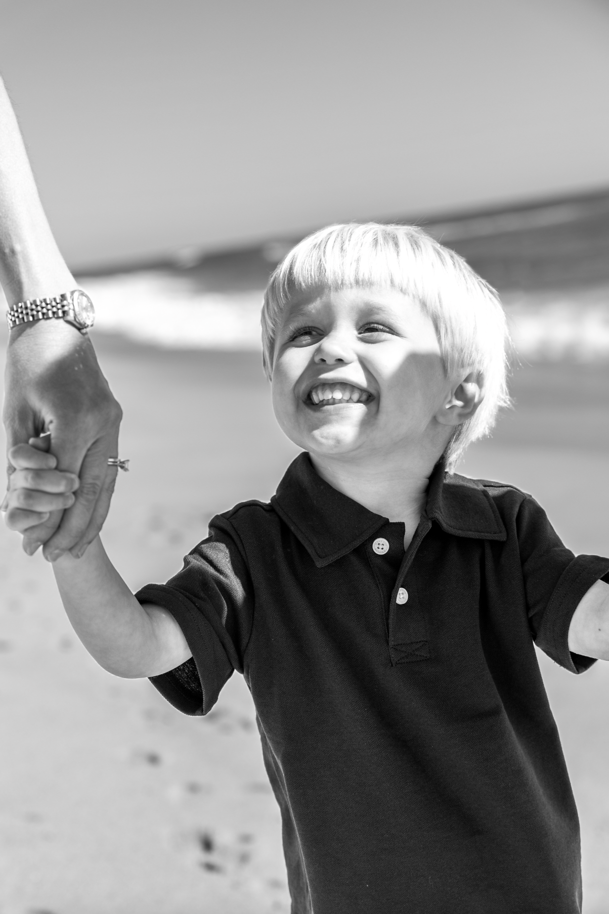 A young boy laughing and smiling while holding hands with an adult on a beach, with the ocean in the background.  Beach Family Photography Monmouth County, New Jersey