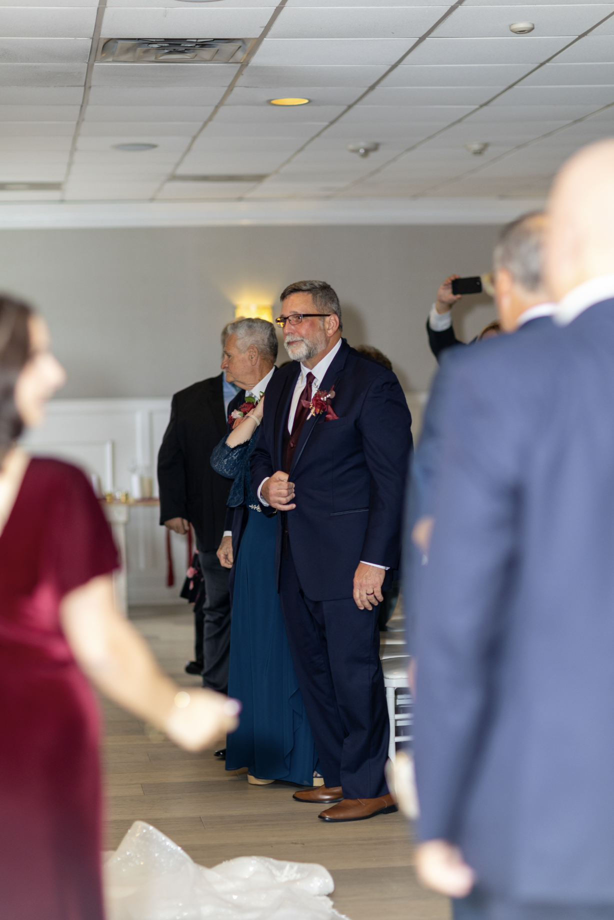 A man in a navy blue suit with a maroon tie and glasses stands with a hand on his chest at a formal event, surrounded by other well-dressed guests, with some taking photos.