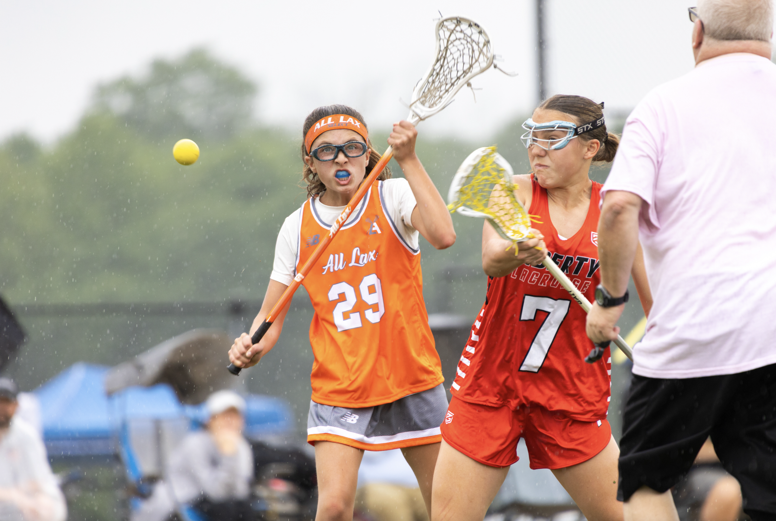 Two female lacrosse players, one wearing an orange jersey with number 29 and the other wearing a red jersey with number 7, are engaged in a game. They are holding lacrosse sticks and are near a referee dressed in a white shirt, with a yellow lacrosse ball in the air between them. It appears to be a competitive match outdoors during a rain shower.