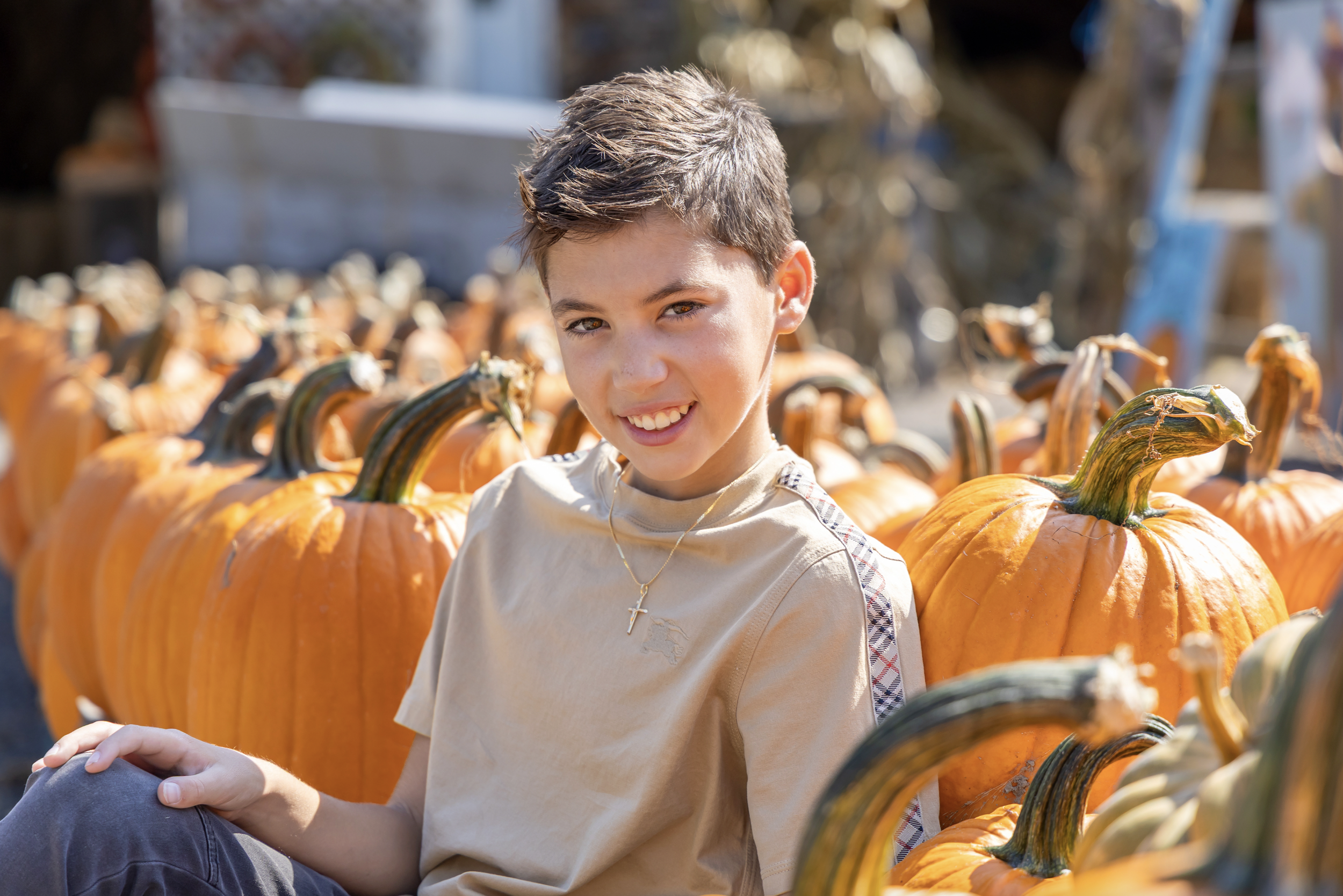 A young boy sitting among pumpkins at a pumpkin patch during daytime, smiling at the camera. Central New Jersey Photography
