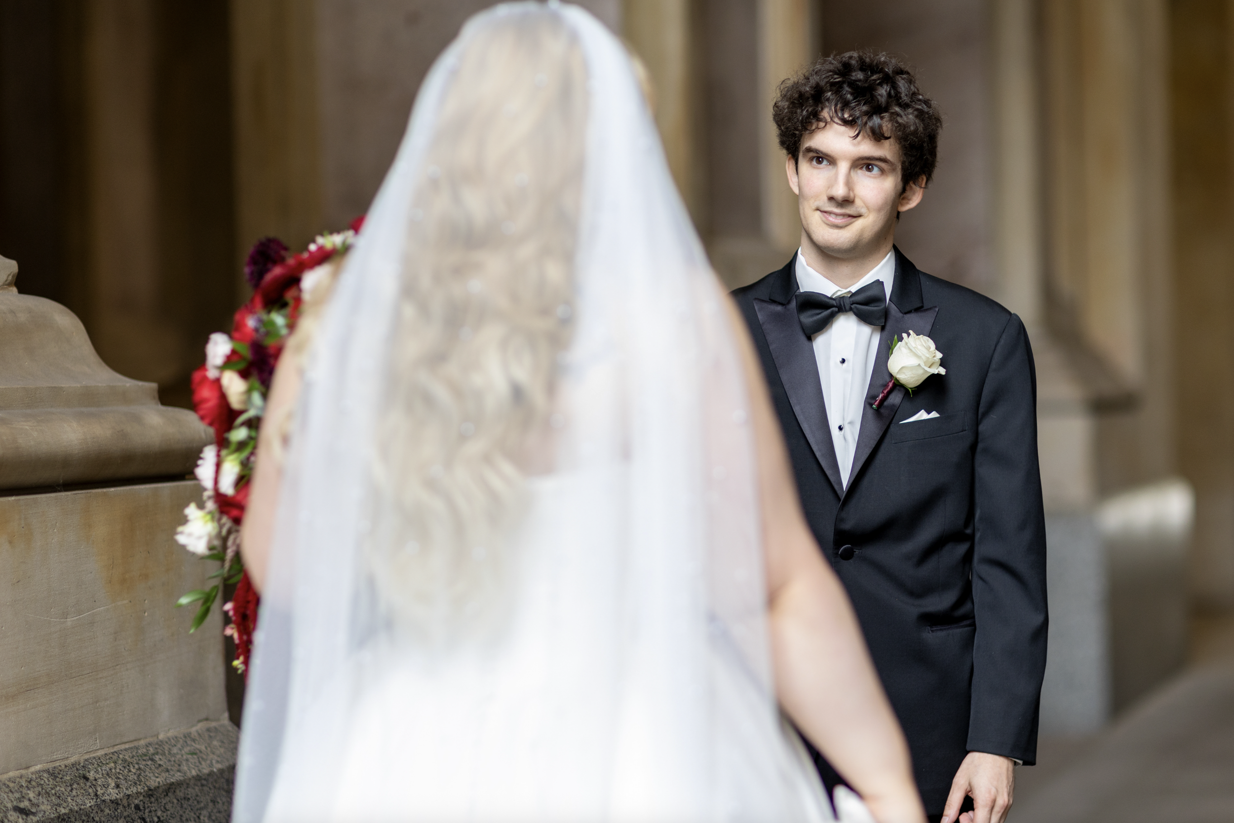 A groom in a black tuxedo with a white boutonniere smiles as he looks at a bride in a wedding dress with a veil and a floral bouquet, inside a grand building with stone architecture.