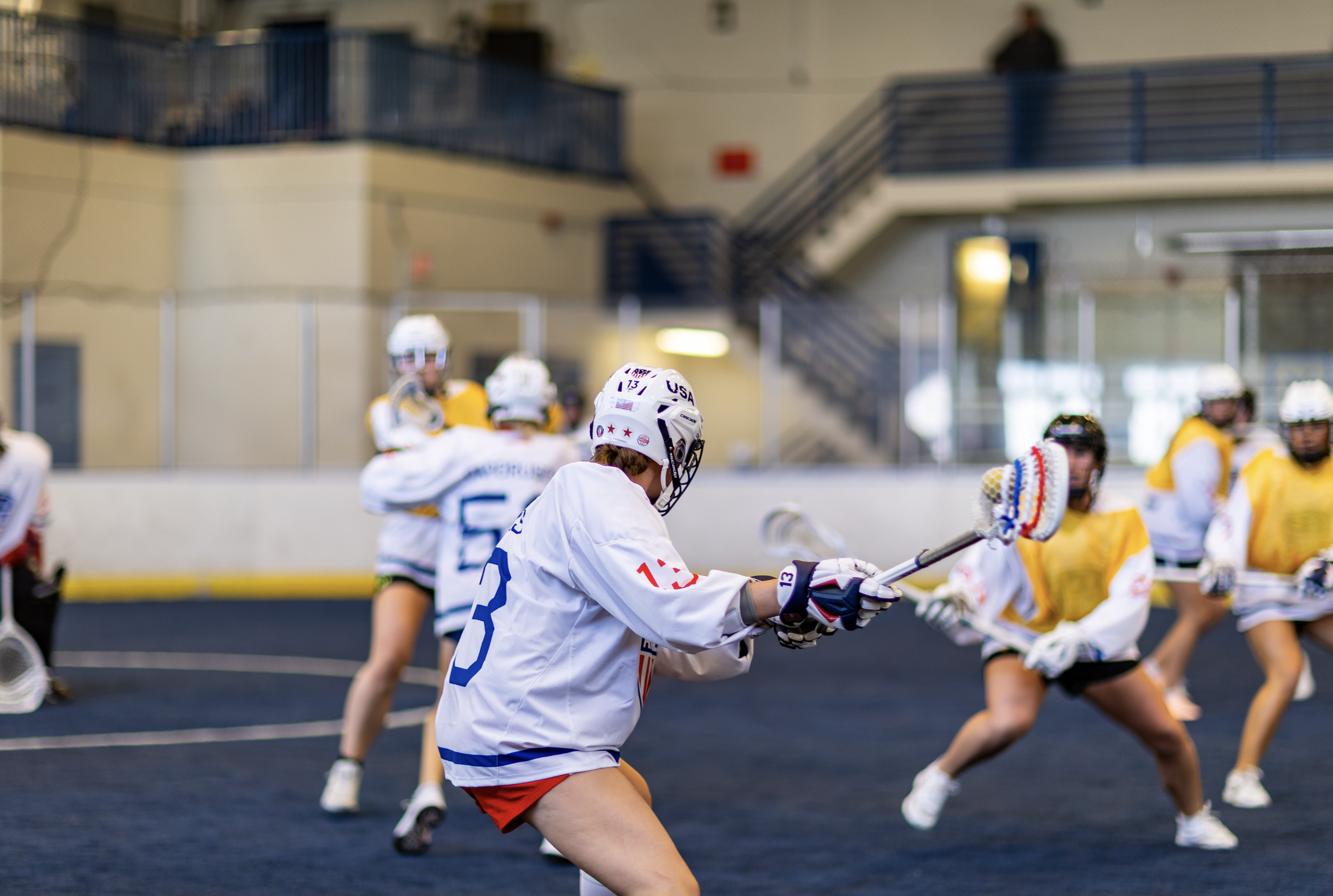 Lacrosse players in action during a game, with one player in a white jersey holding a lacrosse stick preparing to defend or attack, inside an indoor sports facility.