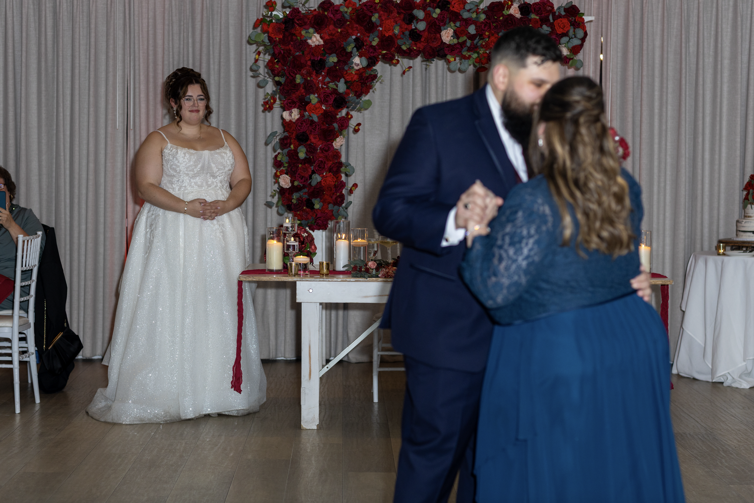 A couple dances closely at their wedding reception, with a large red and pink floral heart backdrop behind them. In the background, a woman in a white wedding gown watches, standing in front of a table with candles and floral decorations. Guests are seated nearby, and a cake is visible on a side table.