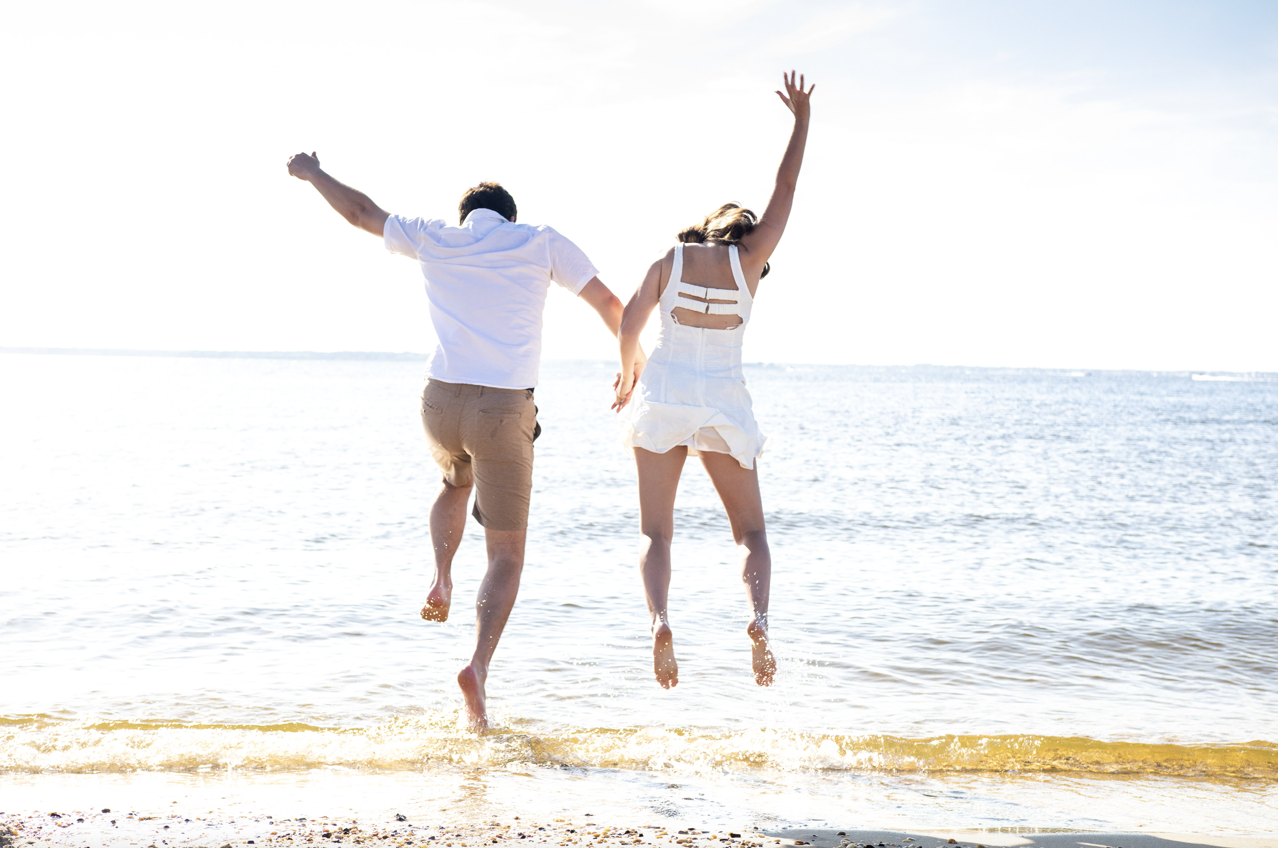 A couple jumping and holding hands at the beach, with water and sky in the background. Ocean County, New Jersey Photographer