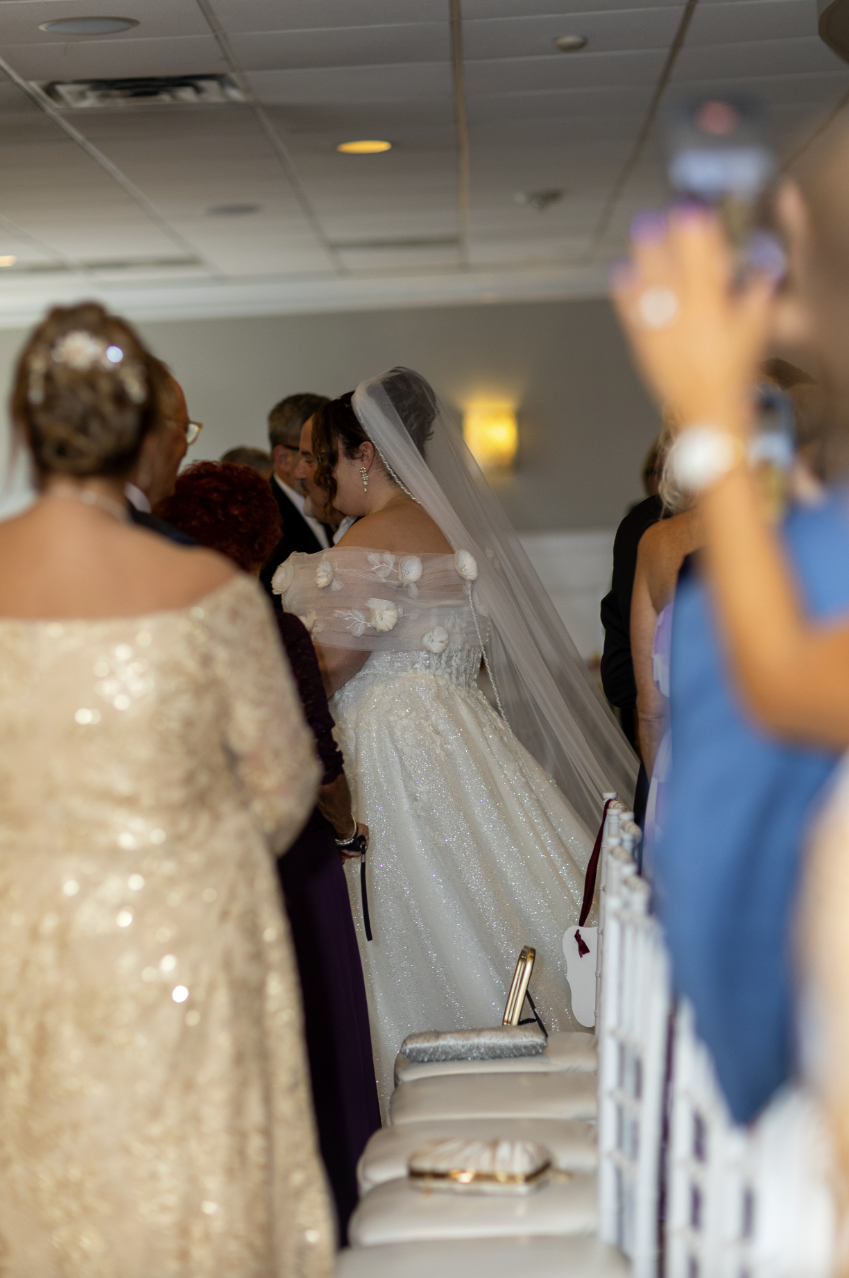 Bride in a white wedding gown with a veil and floral details, surrounded by wedding guests at an indoor celebration.