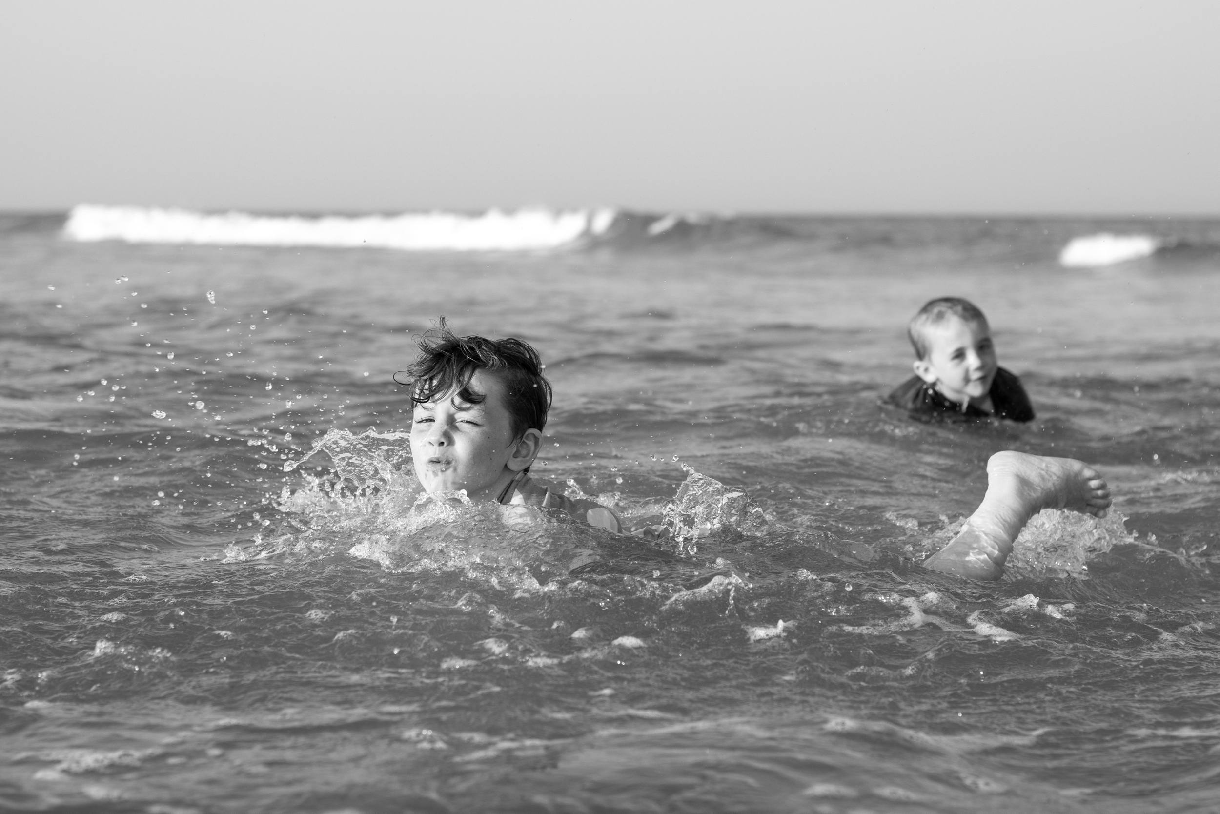 Two children playing in the ocean, one splashing water and the other swimming, with waves in the background.