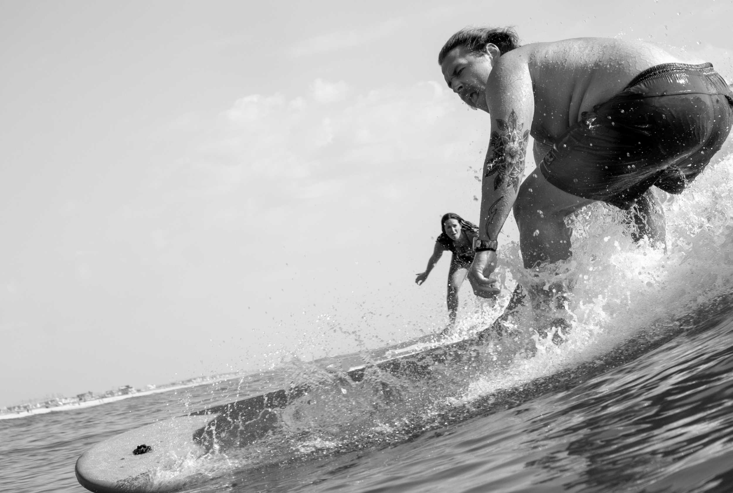 Two people surfing in the ocean, a man with tattoos on his arm in the foreground and a woman behind him in the background.