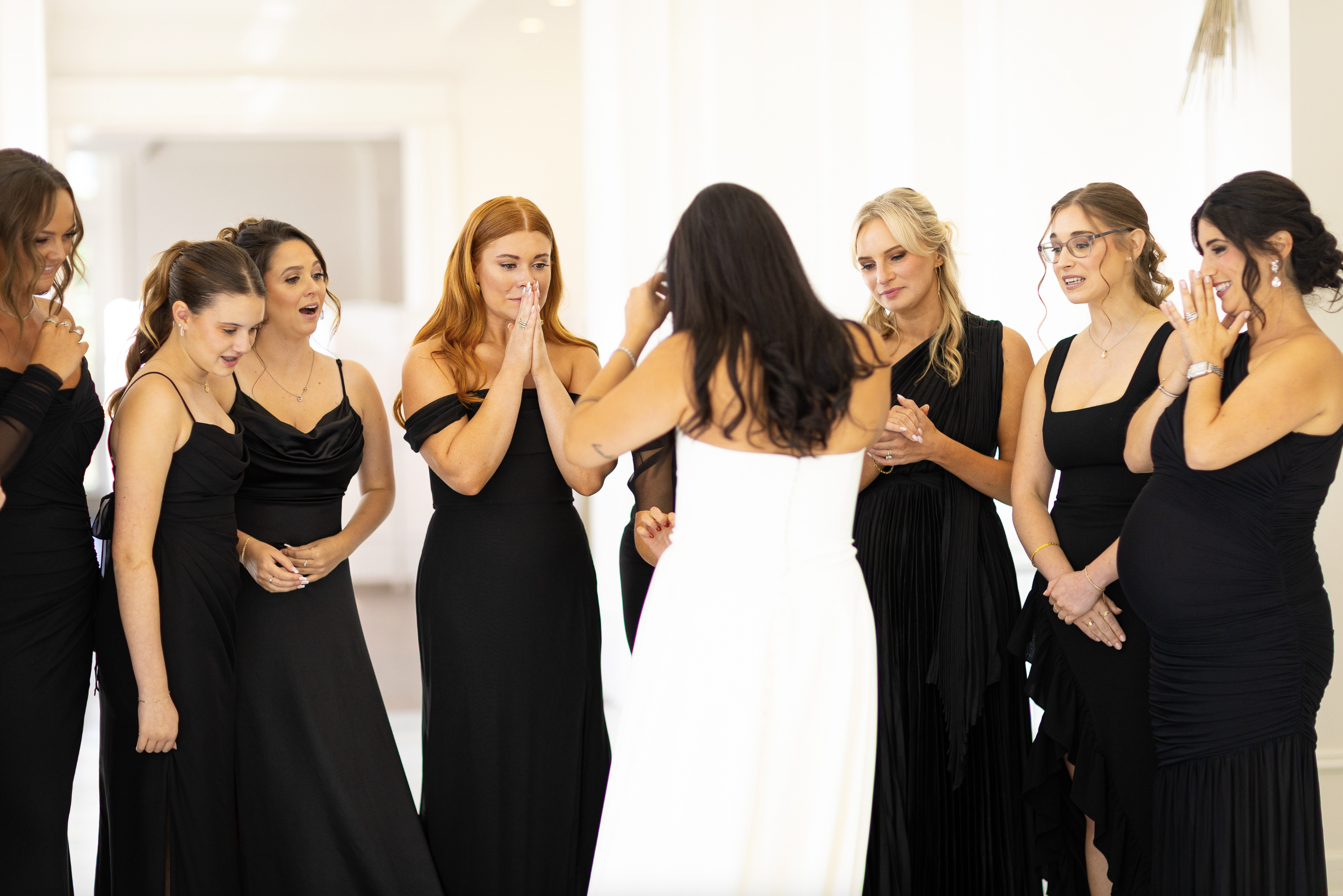 A bride in a white dress surrounded by eight women in black dresses, all reacting emotionally during a wedding moment.