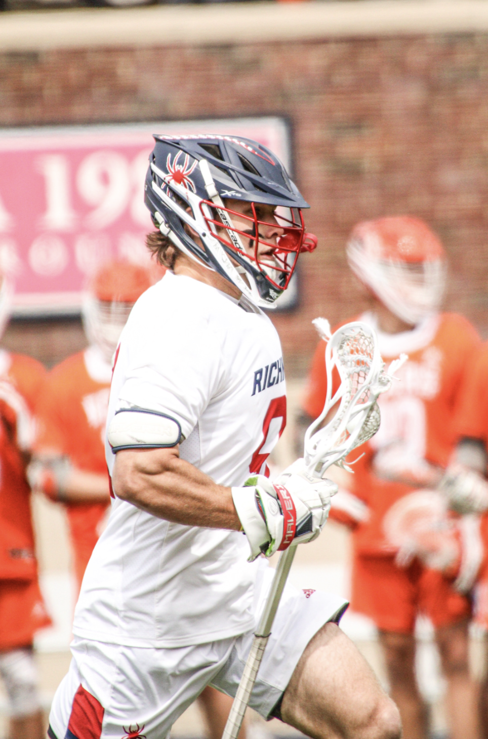 A male lacrosse player wearing a Richmond uniform and helmet, holding a lacrosse stick, running on the field with teammates in orange uniforms in the background.
