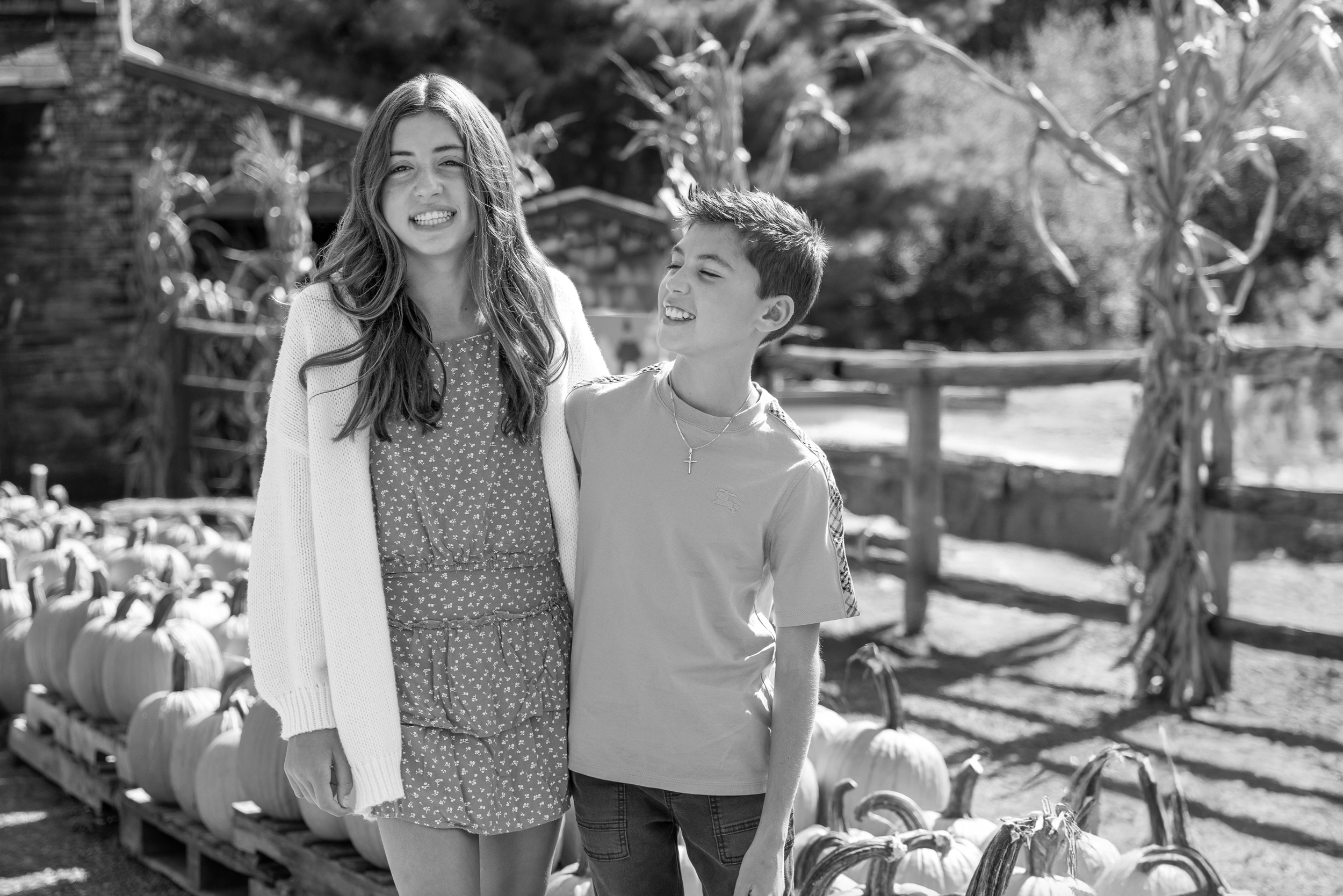 A girl and boy smiling and standing close together outdoors at a pumpkin patch, with pumpkins on the ground and a rustic wooden fence in the background.
