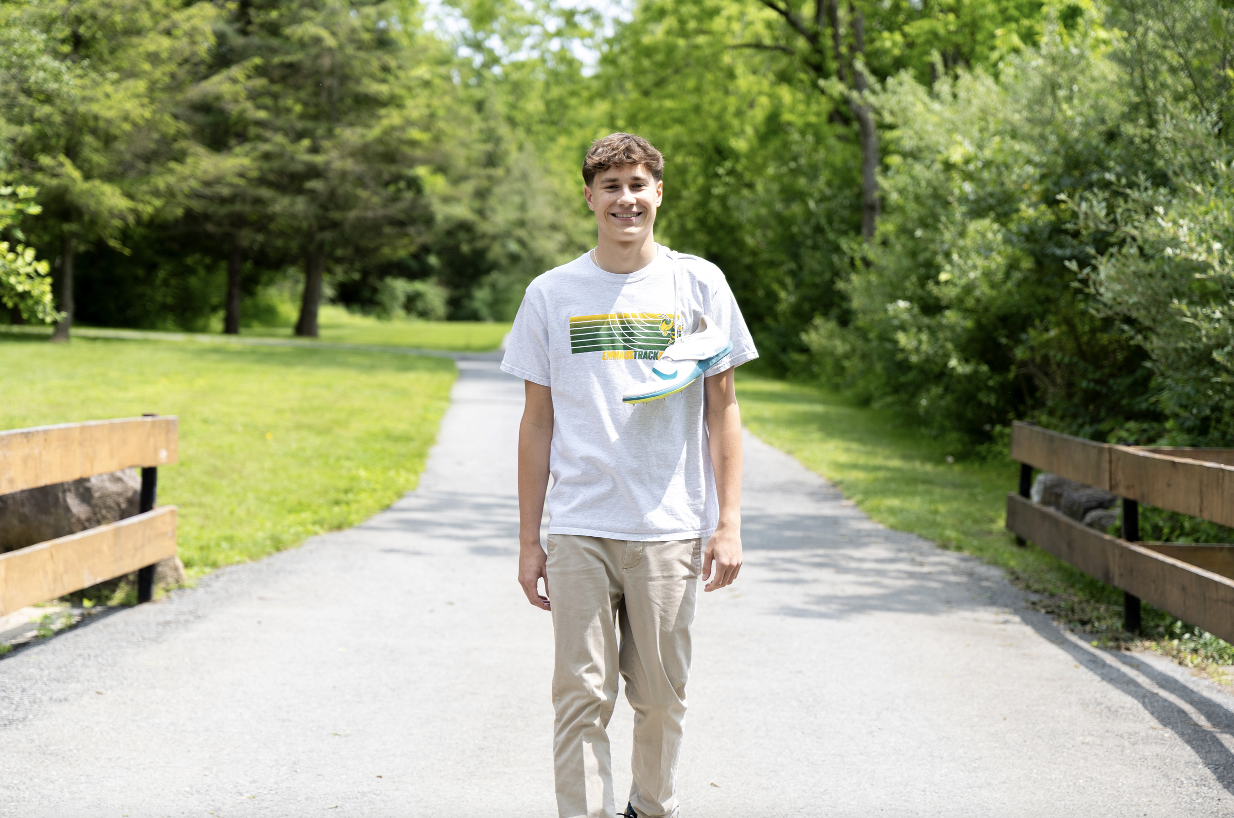 A young man walking on a paved trail in a park with lush green trees and grass, smiling at the camera. Pennsylvania, Bucks County high school senior pictures