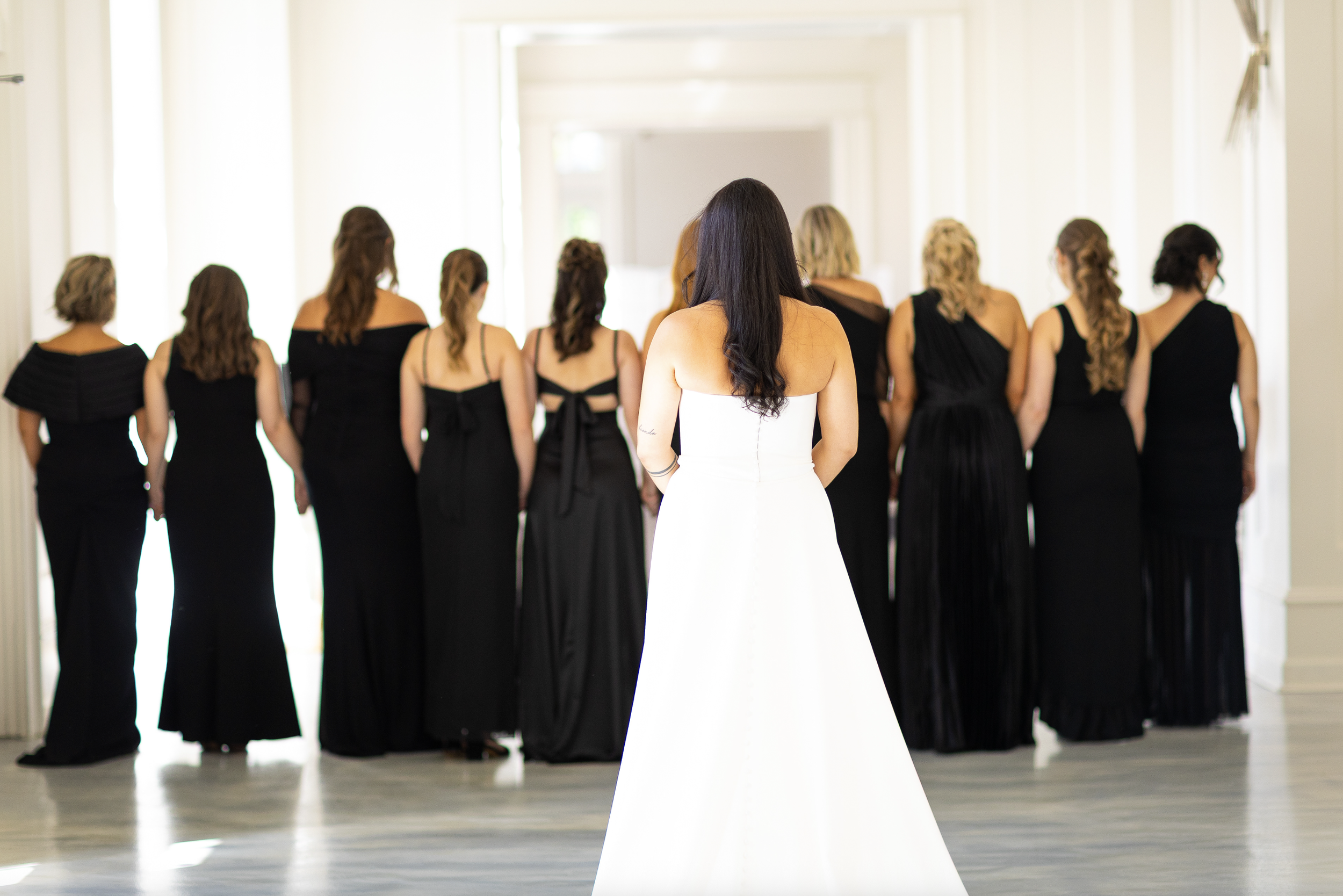 A bride in a white wedding dress watches her bridesmaids in black dresses as they walk in a line in a bright, airy hallway.