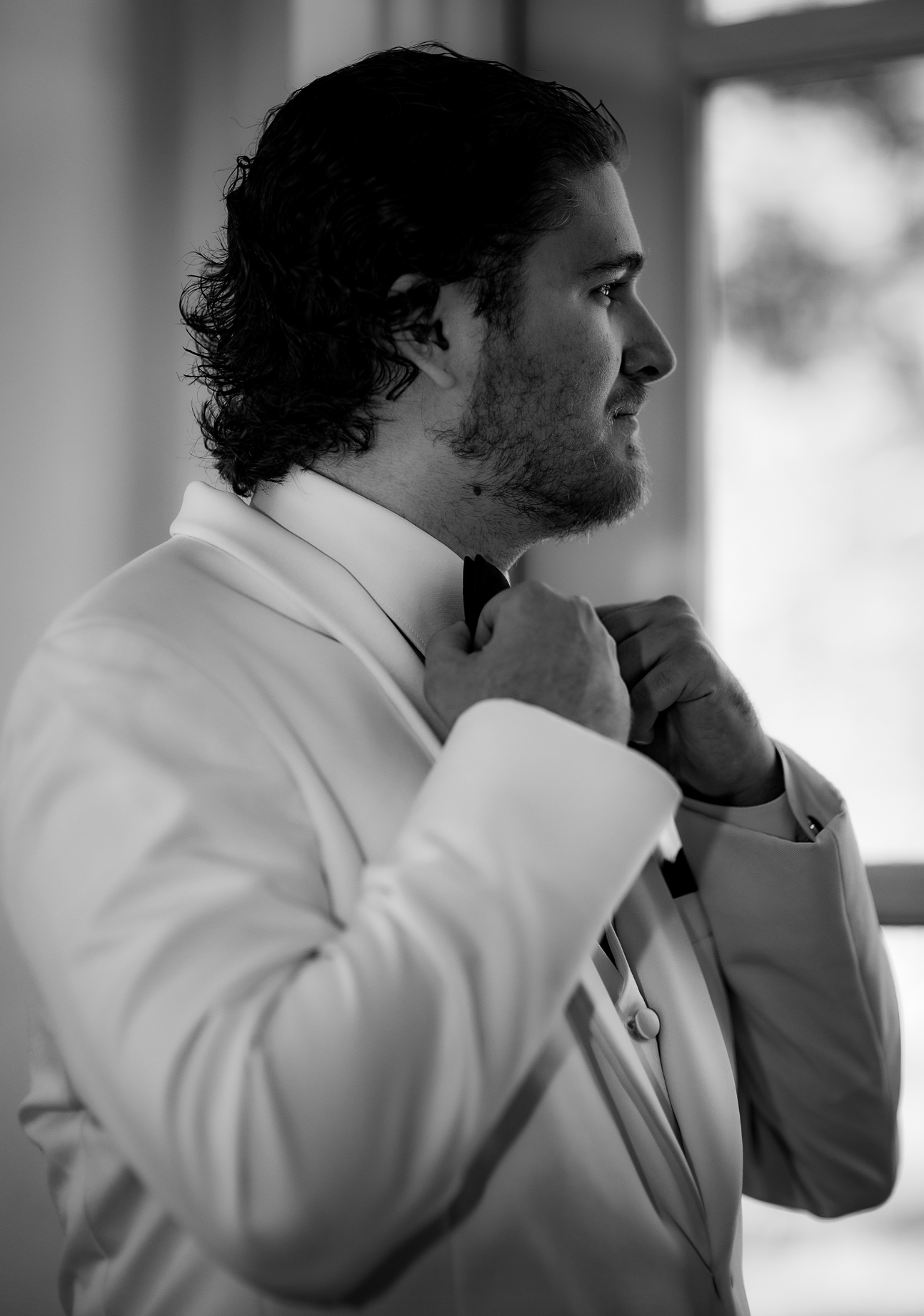 A man in a tuxedo adjusts his bow tie, preparing for a formal event, standing near a window with sunlight coming through. jersey shore groom wedding photography