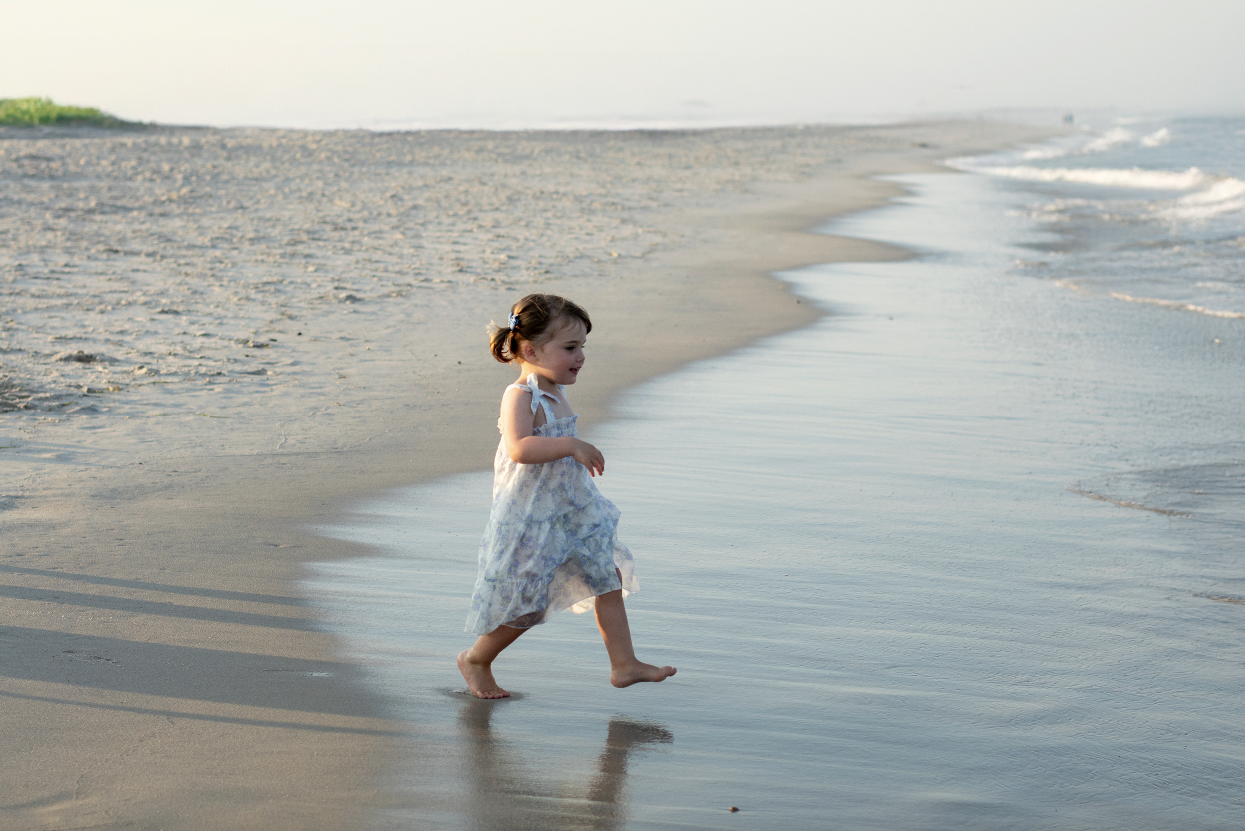 A young girl in a light-colored dress is running barefoot along the shoreline of a beach, with waves touching her feet and a green patch of land in the background.  Beach Family Photography New Jersey