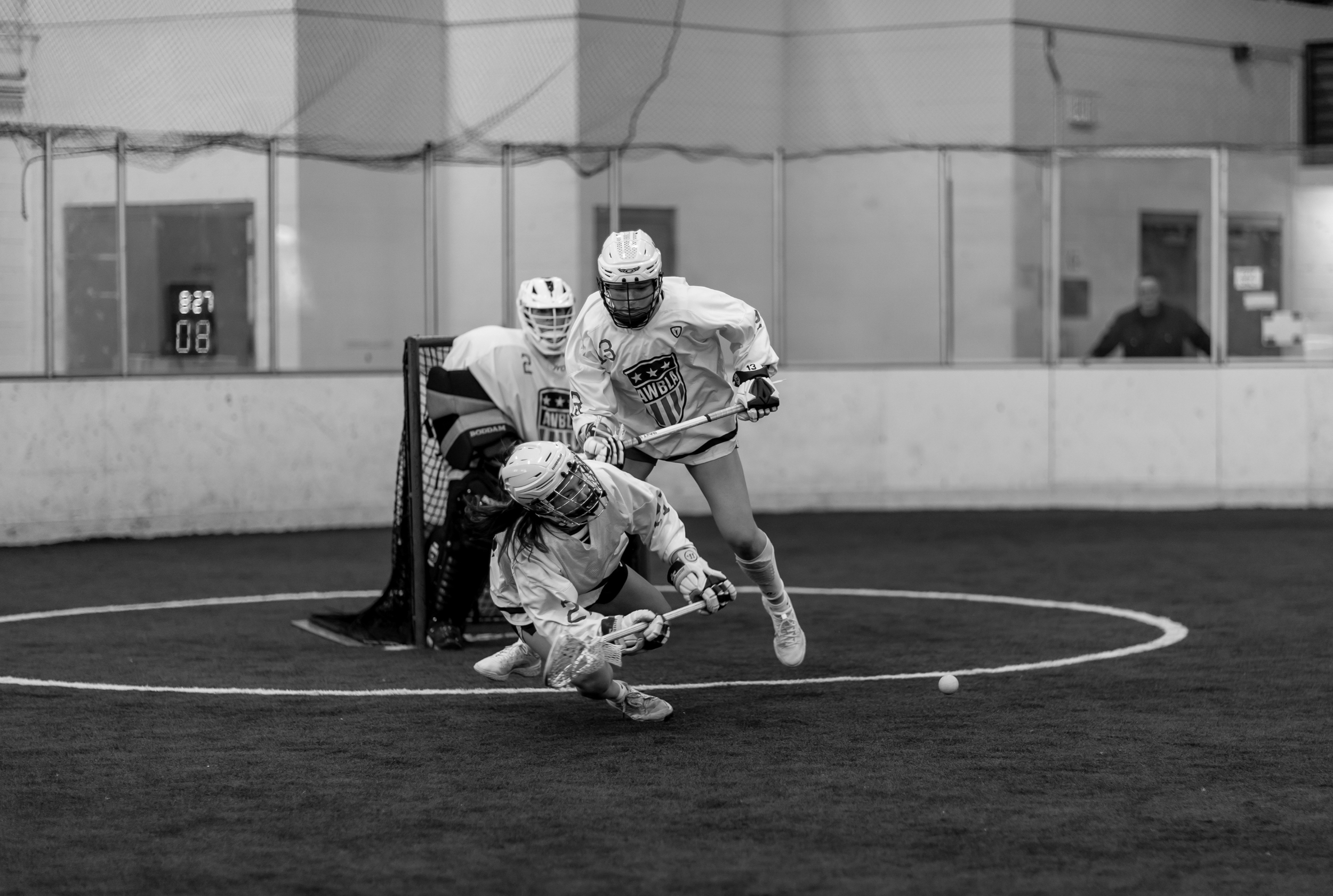 Three girls playing lacrosse inside an indoor sports facility, with one girl falling to the ground and the other two attempting to gain possession of the ball near the goal.