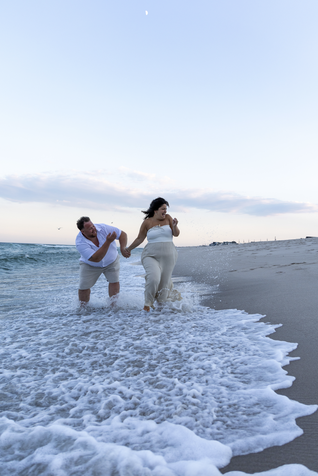 A man and woman holding hands and laughing while walking into the ocean at sunset or dusk on a beach. Ocean County, New Jersey Engagement Photography
