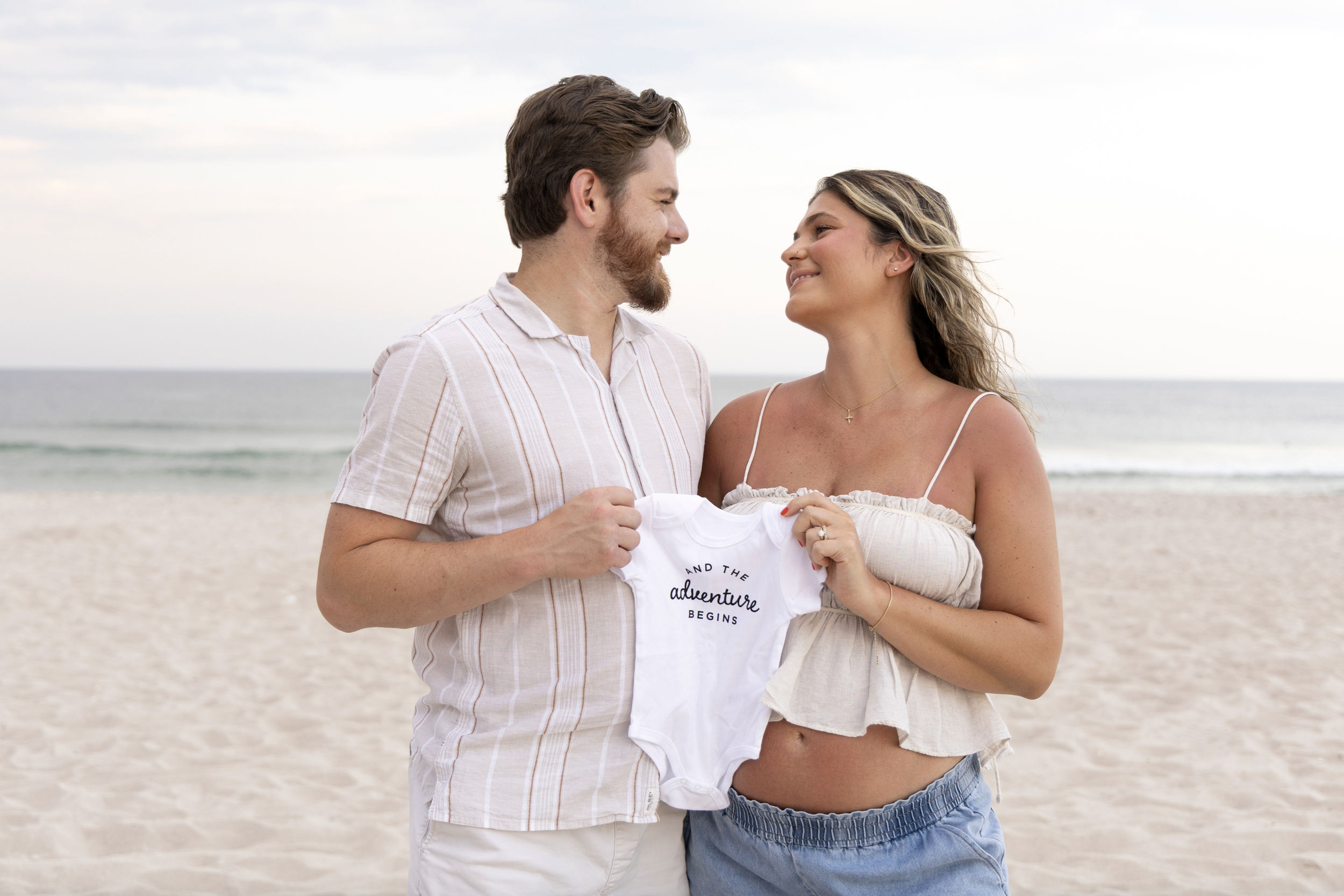 A couple stands on a beach holding a white baby onesie that reads 'and the adventure begins.' They smile at each other with the ocean in the background. Ocean Beach, New Jersey