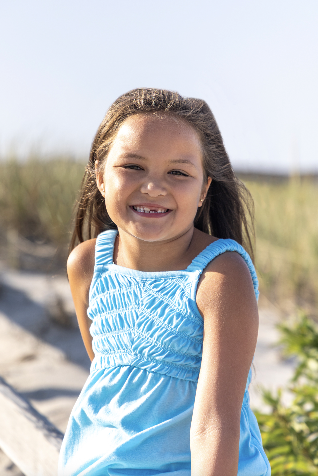 A smiling young girl with brown hair wearing a light blue dress at the beach. Beach Family Photography Ocean County, New Jersey