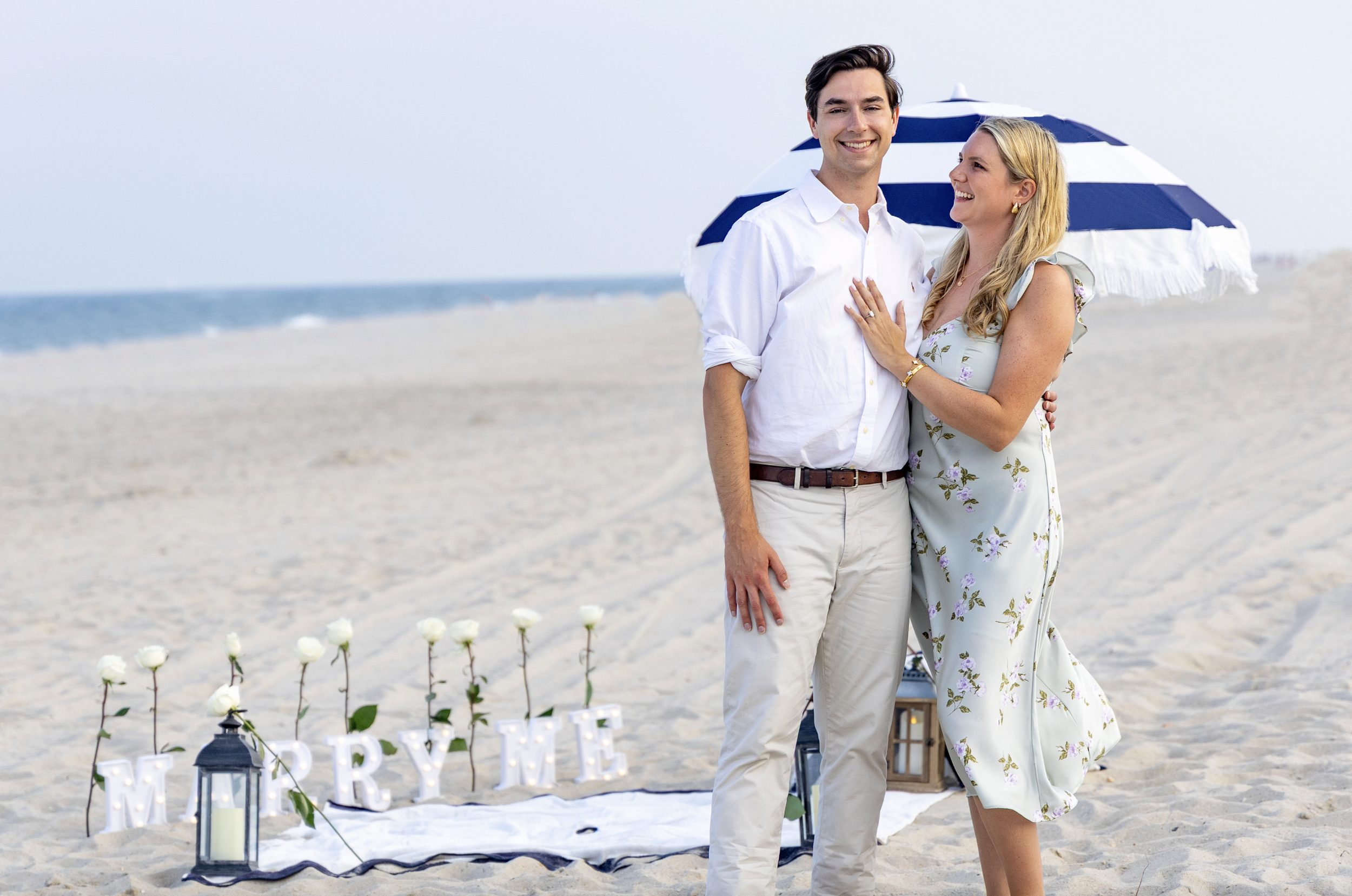 A couple on the beach with a small wedding setup, including white roses, lanterns, and illuminated "MARRY ME" letters, under a striped beach umbrella. Lavallette, Ocean County, NJ