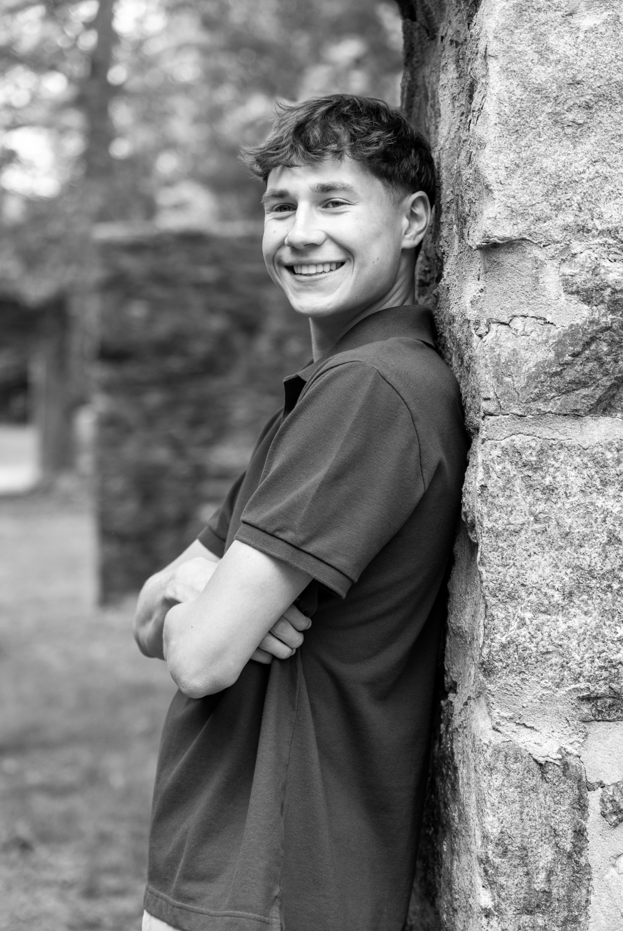 A smiling young man leaning against a stone wall outdoors.