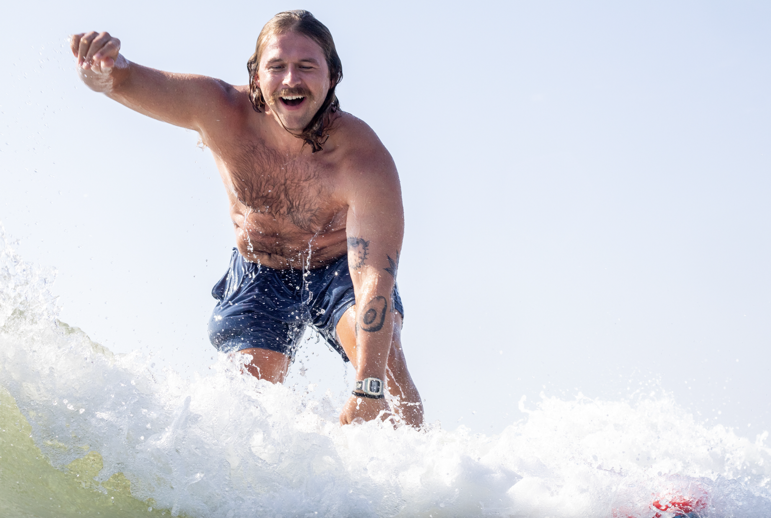 A man surfing on a wave, smiling with wet hair, wearing dark swim trunks, with tattoos on his arm and a wristwatch.
