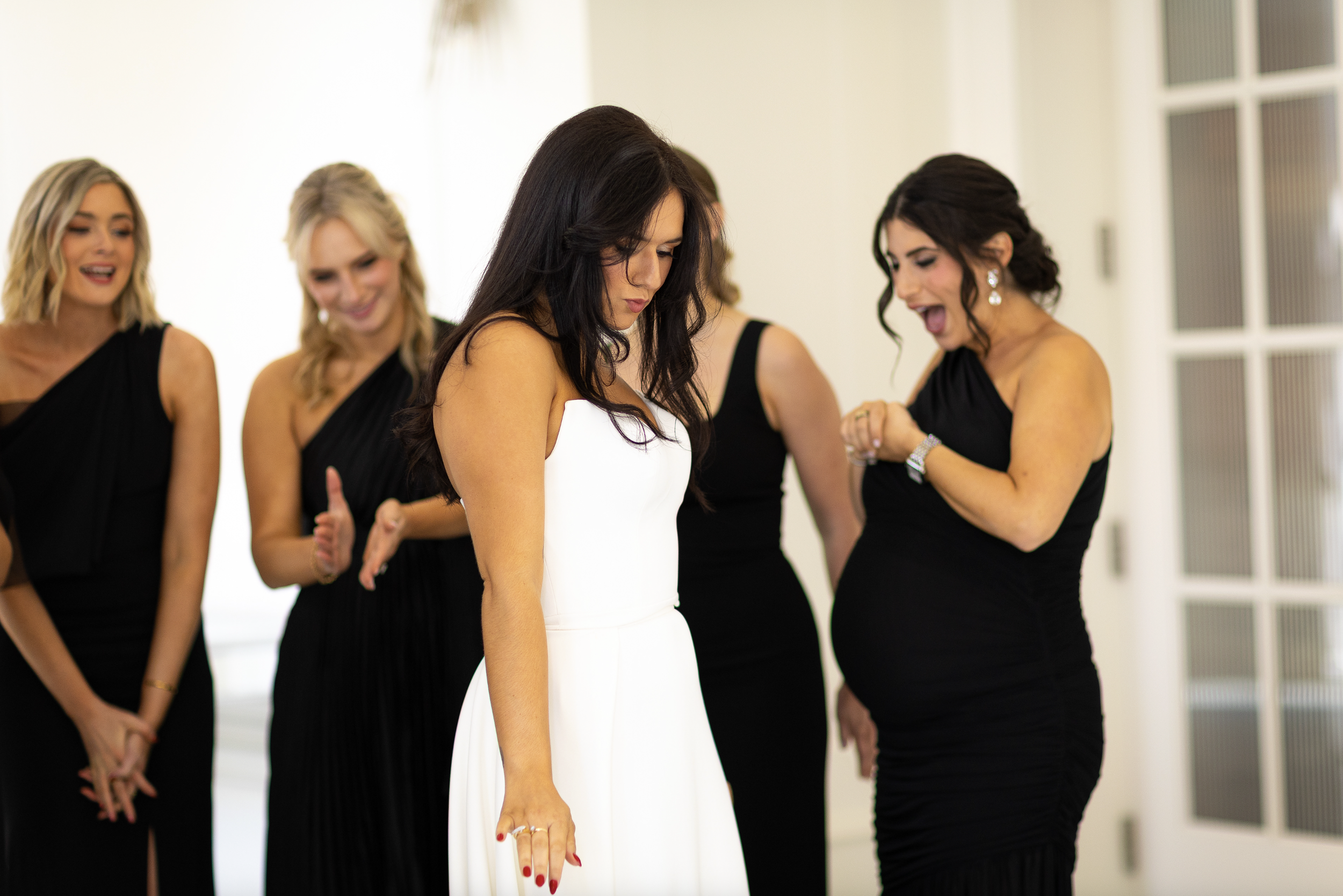 Women in black dresses and one in a white dress gathered indoors, appearing to prepare the bride for her wedding.