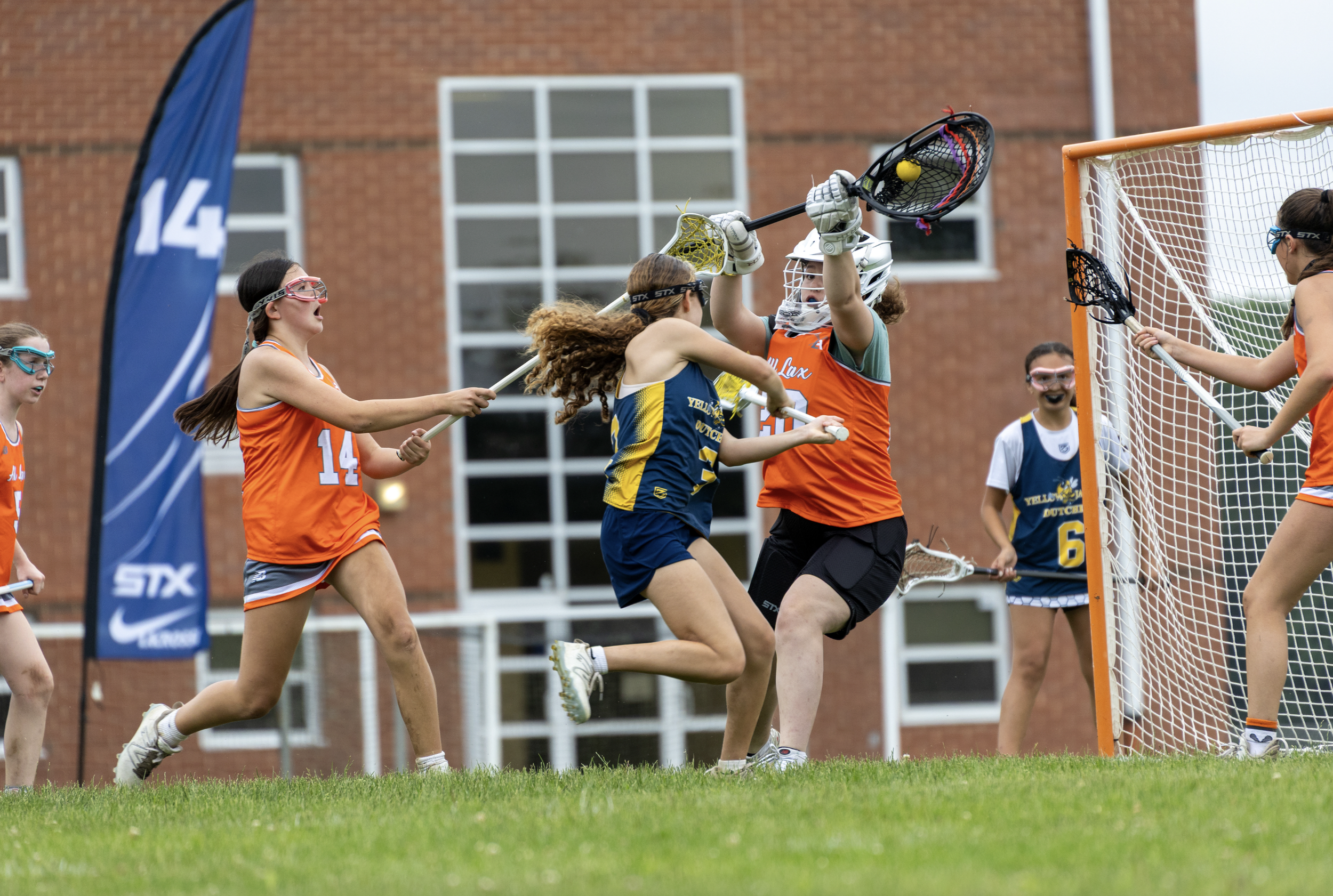 Girls playing lacrosse near the goal, with one goalie attempting to block a shot while others chase the ball.