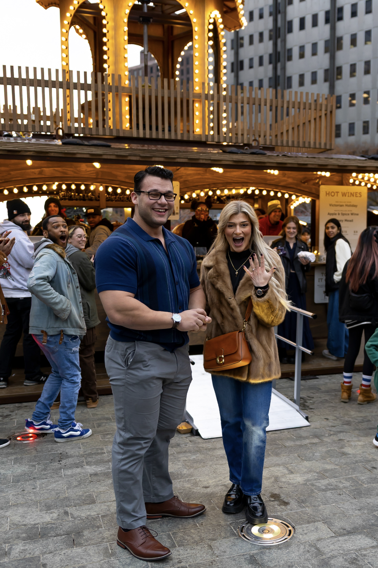 People at a festive outdoor event, with a lit-up carousel in the background, some smiling and others cheering, on a paved city street. Love Park, Philadelphia surprise proposal photography