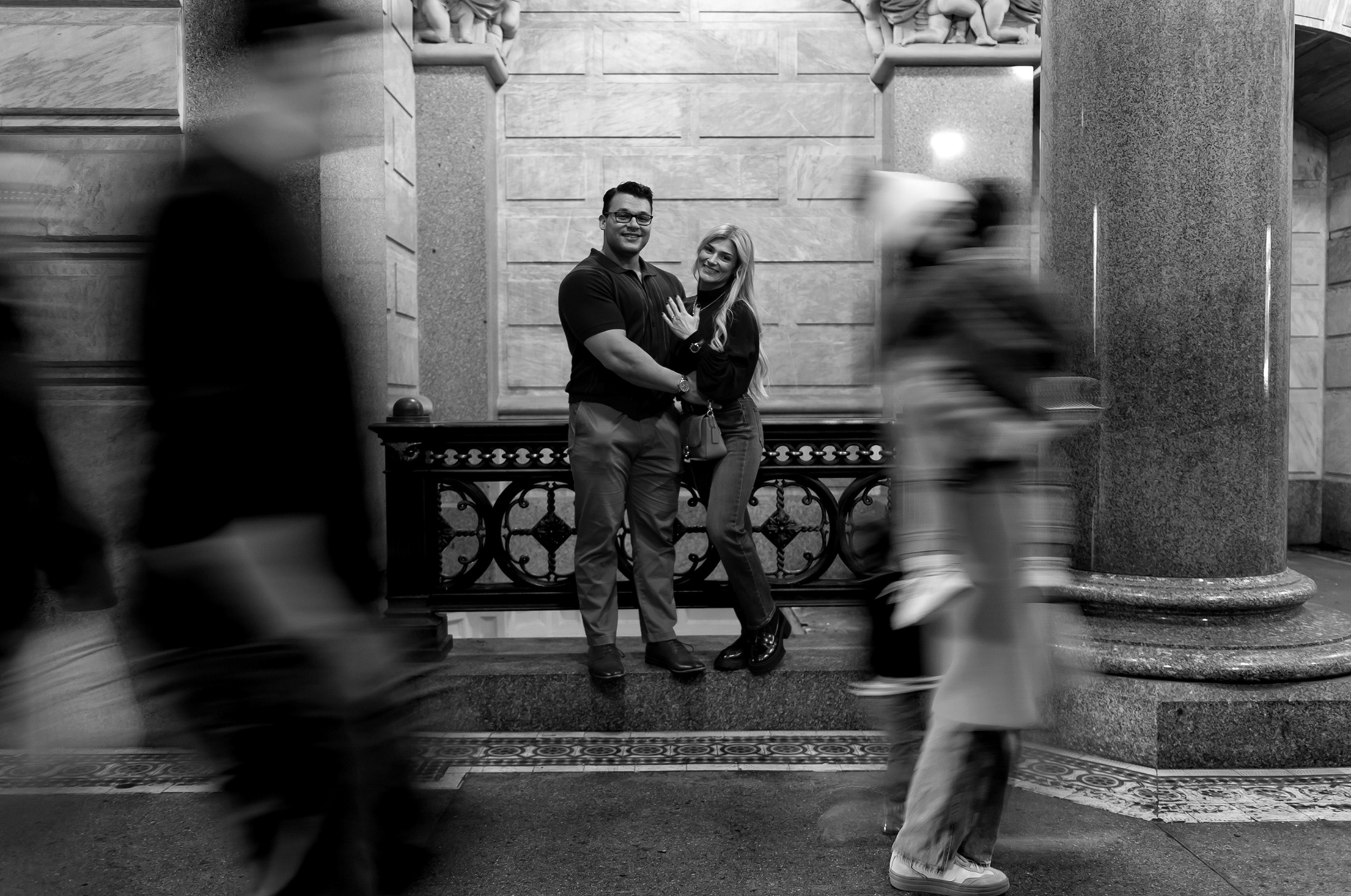 A couple stands close together, smiling, in a busy indoor setting with blurred pedestrians walking past. The man is wearing glasses and a dark shirt, and the woman has long blonde hair and is wearing a dark top and jeans. The background features large stone columns and artistic elements.
